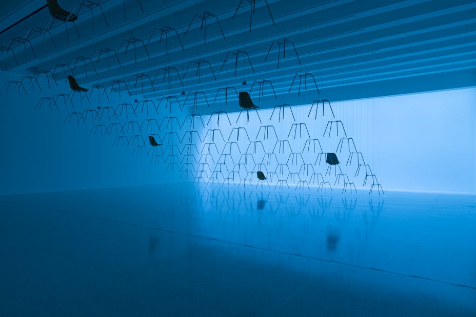 Gallery bathed in blue light with chairs and chair legs suspended from the ceiling