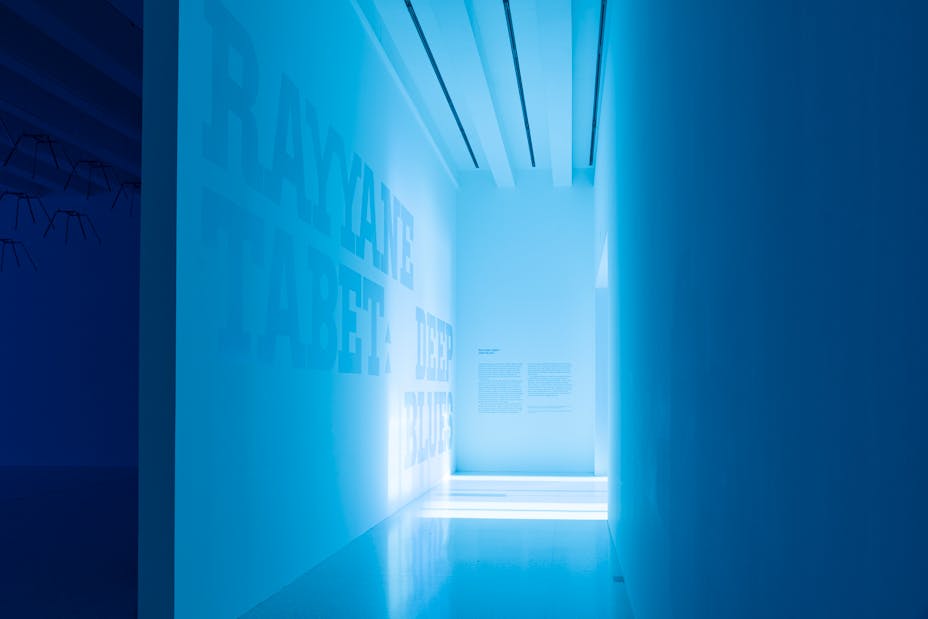 Entryway to gallery with large scale iridescent typography on wall that says "Rayyane Tabet: Deep Blues"