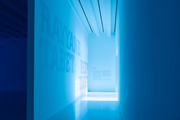 Entryway to gallery with large scale iridescent typography on wall that says "Rayyane Tabet: Deep Blues"