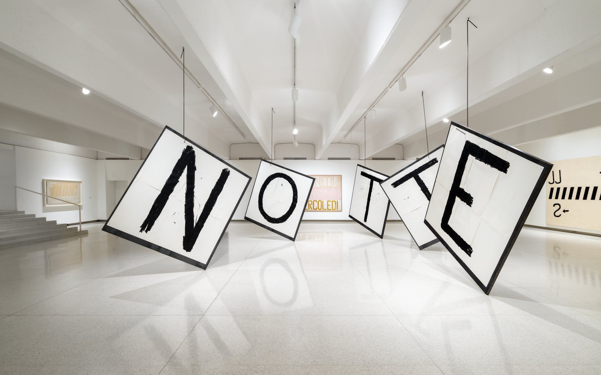 A series of paintings of single letters are hung by large metal hooks in a gallery.