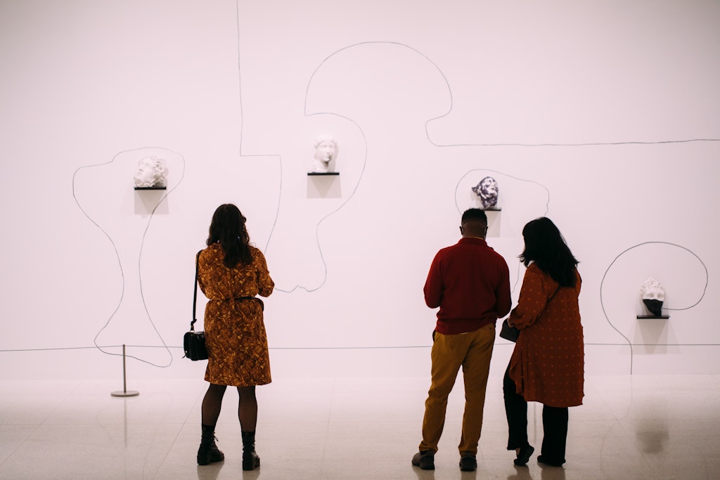 Three people stand in front of an artwork of lines drawn on a wall and fragments of classic sculptures.