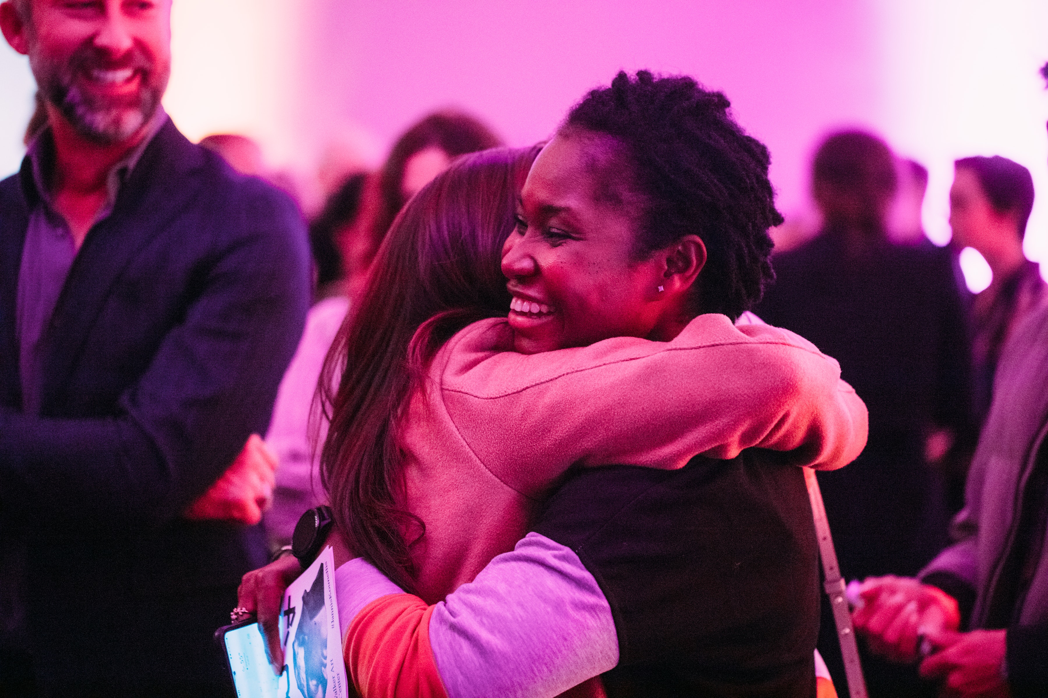 Two adults smile and hug in a crowd bathed in colored lights in a museum lounge.