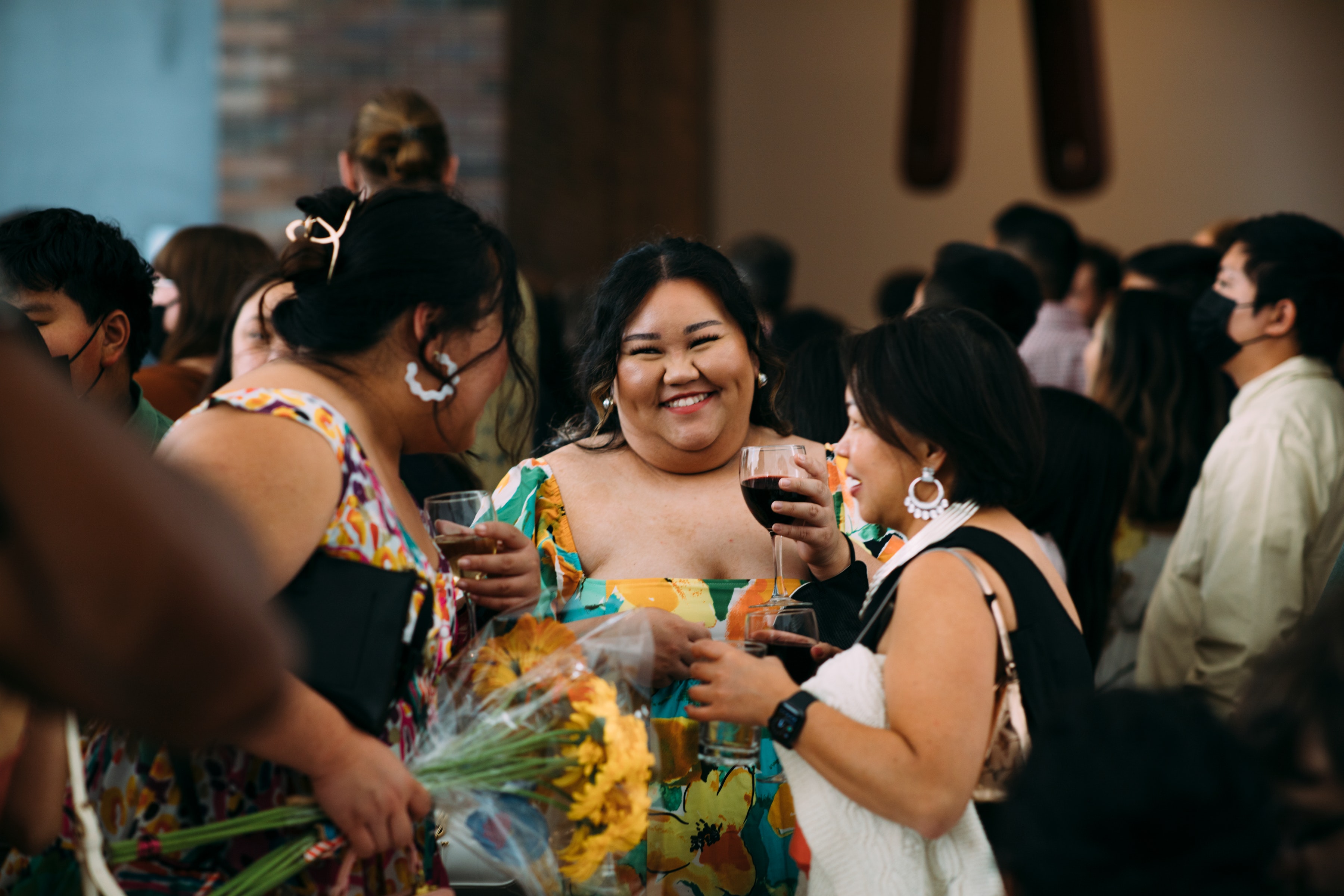 A group of adults wearing colorful dresses smile while holding wine and flowers in a crowd.