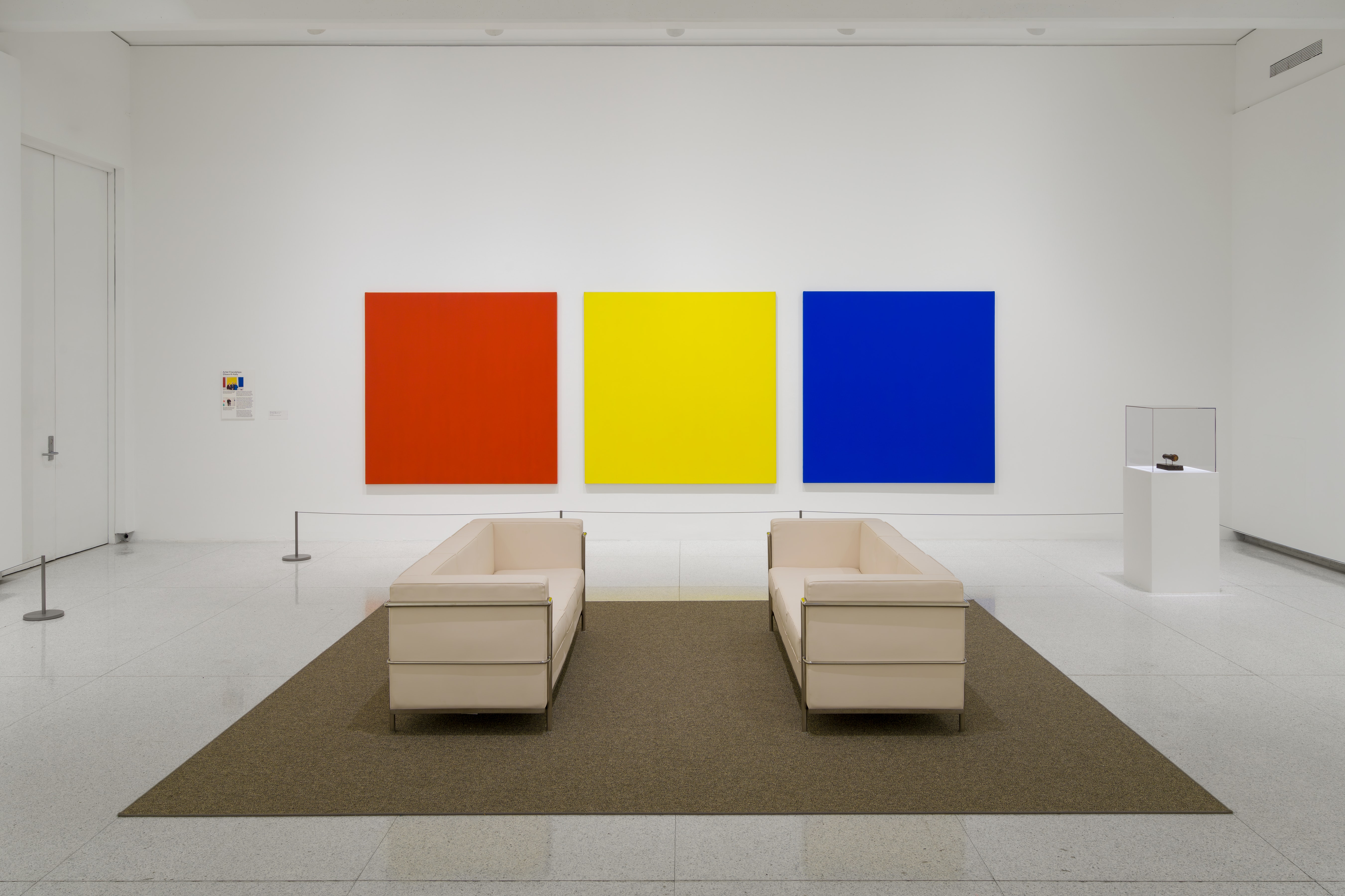 Three solid color paintings, one red, one yellow, and one blue, hang in a white art gallery in front of two white couches.