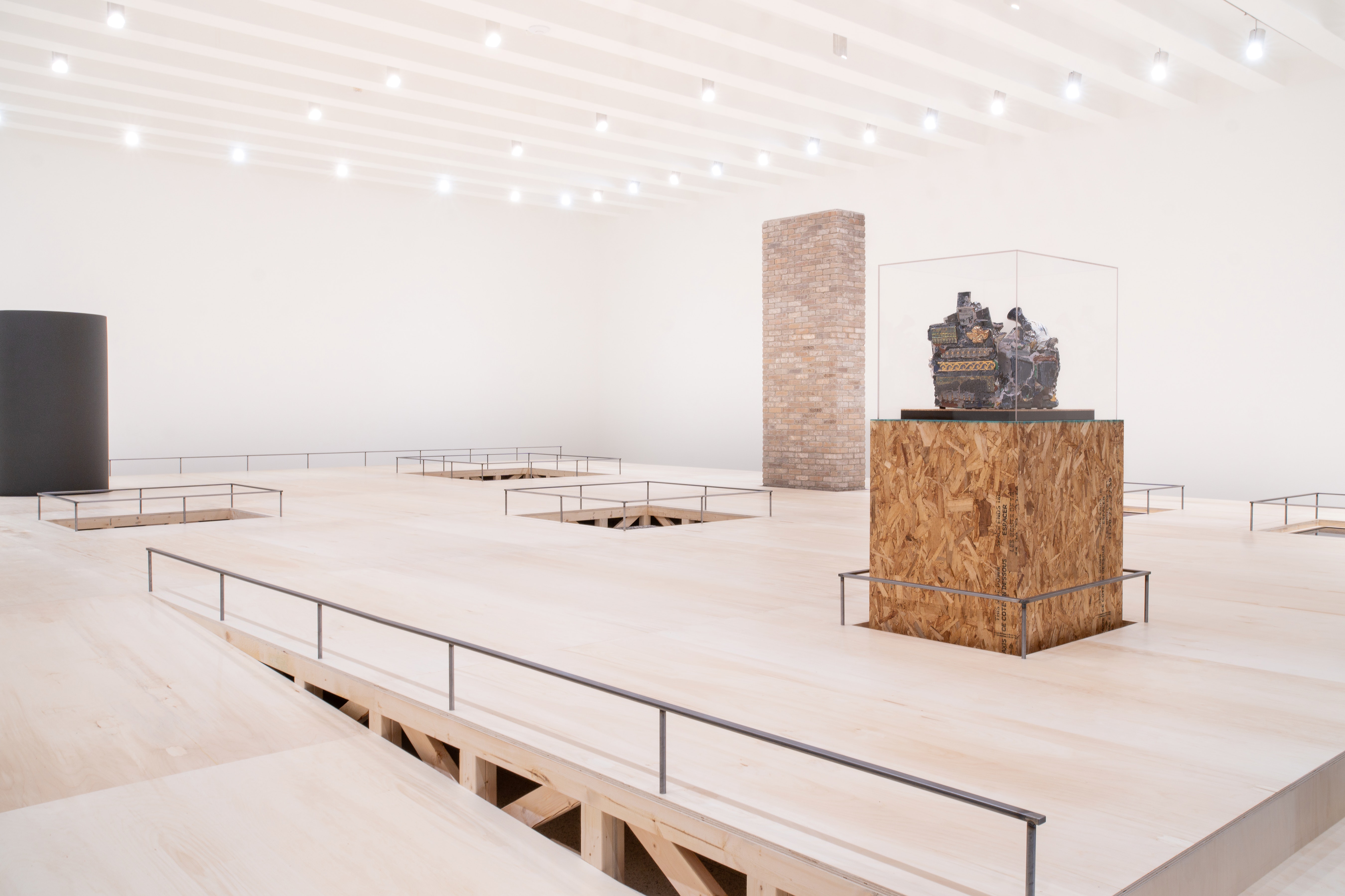 A brick wall, large cermaic tube, and a ceramci artwork on a pedistal rise up out of a raw wood platform with rails and a ramp that spans nearly the entire space of a white box art gallery.