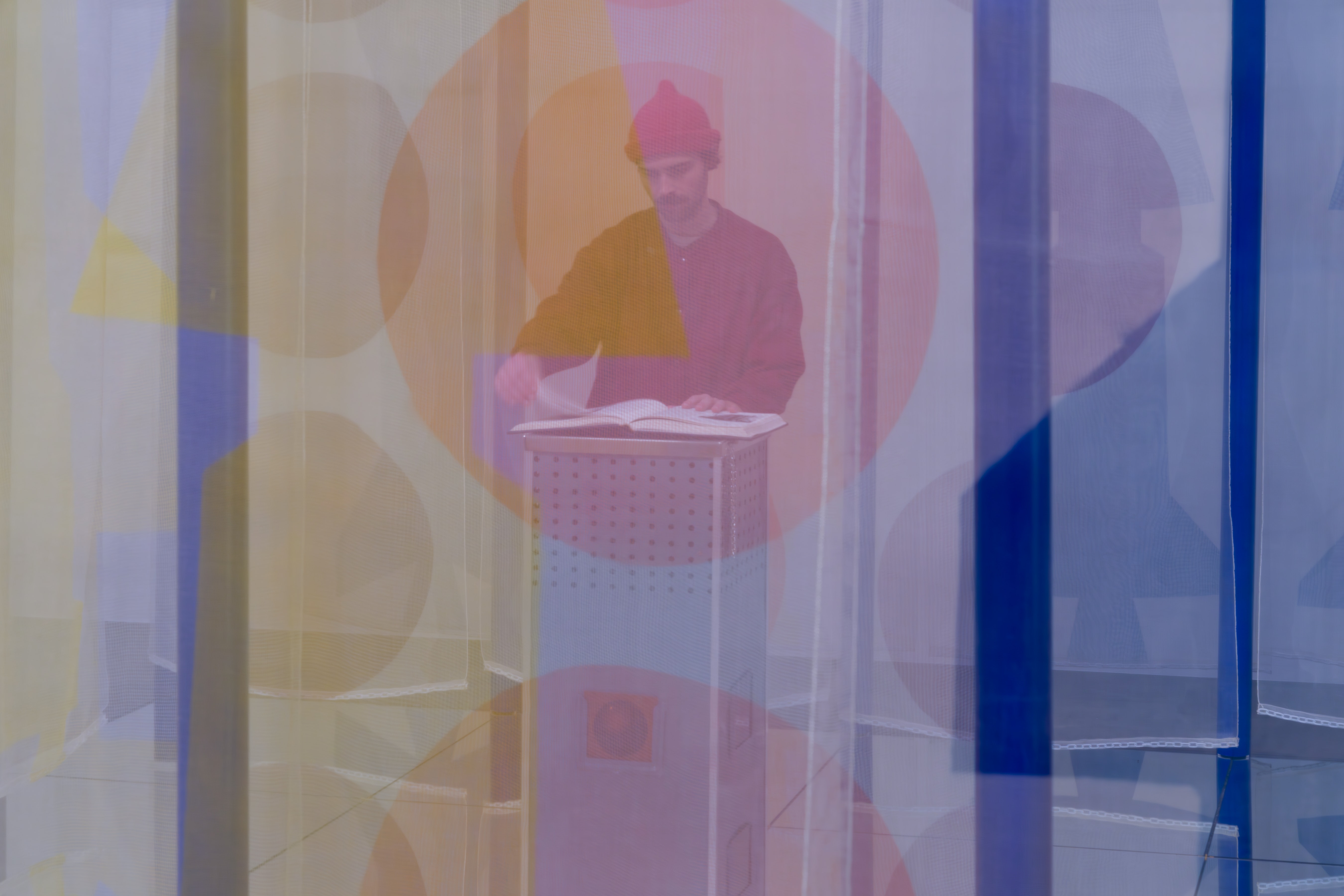 An adult stand inside a exhibit looking at a book and it obscured by a colorful semi-transparent screen.