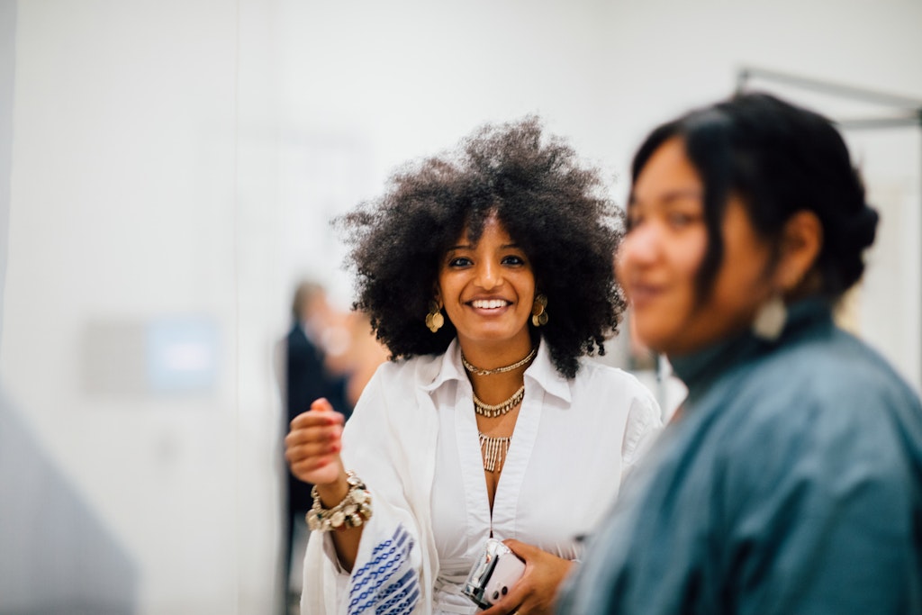 An adult with medium dark skin stands in a gallery space and faces the viewer smiling, a blurred out person in the foreground.