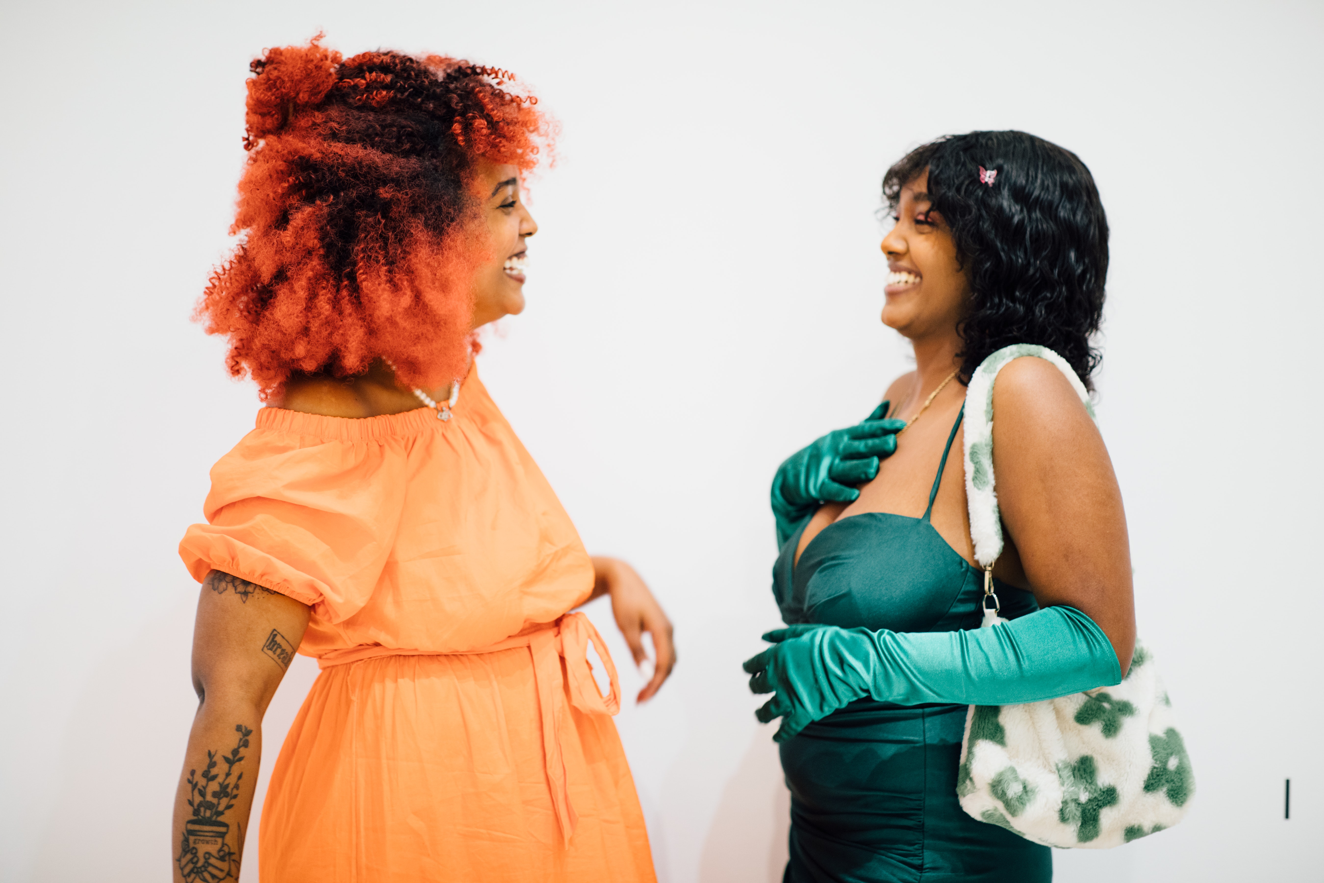 Two medium skinned adults in colorful clothing stand facing eachother, laughing in a white space.