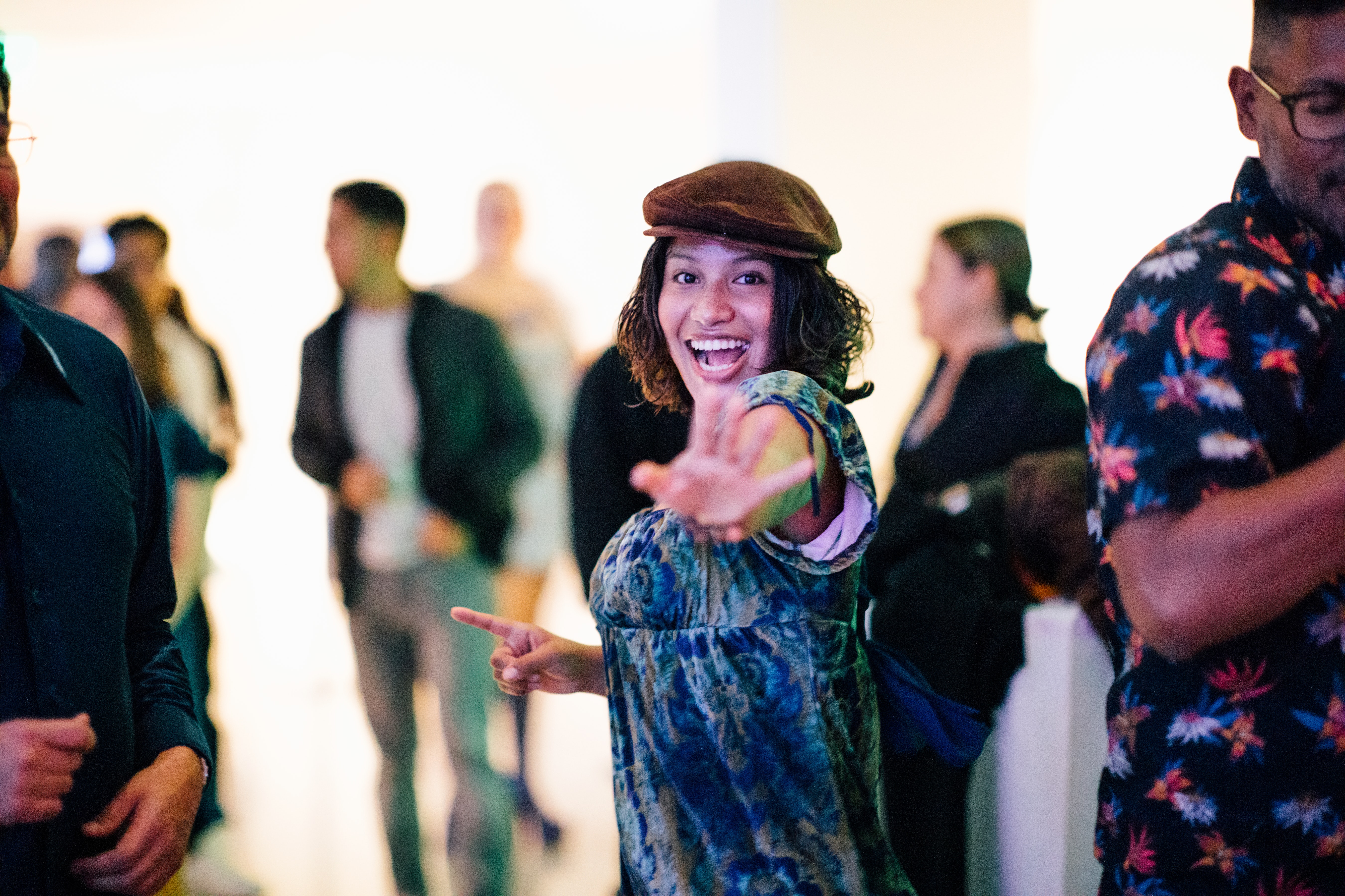 A medium skinned adult wearing a hat and dress smiles and reaches out towards viewer in a blurred-out crowd around
