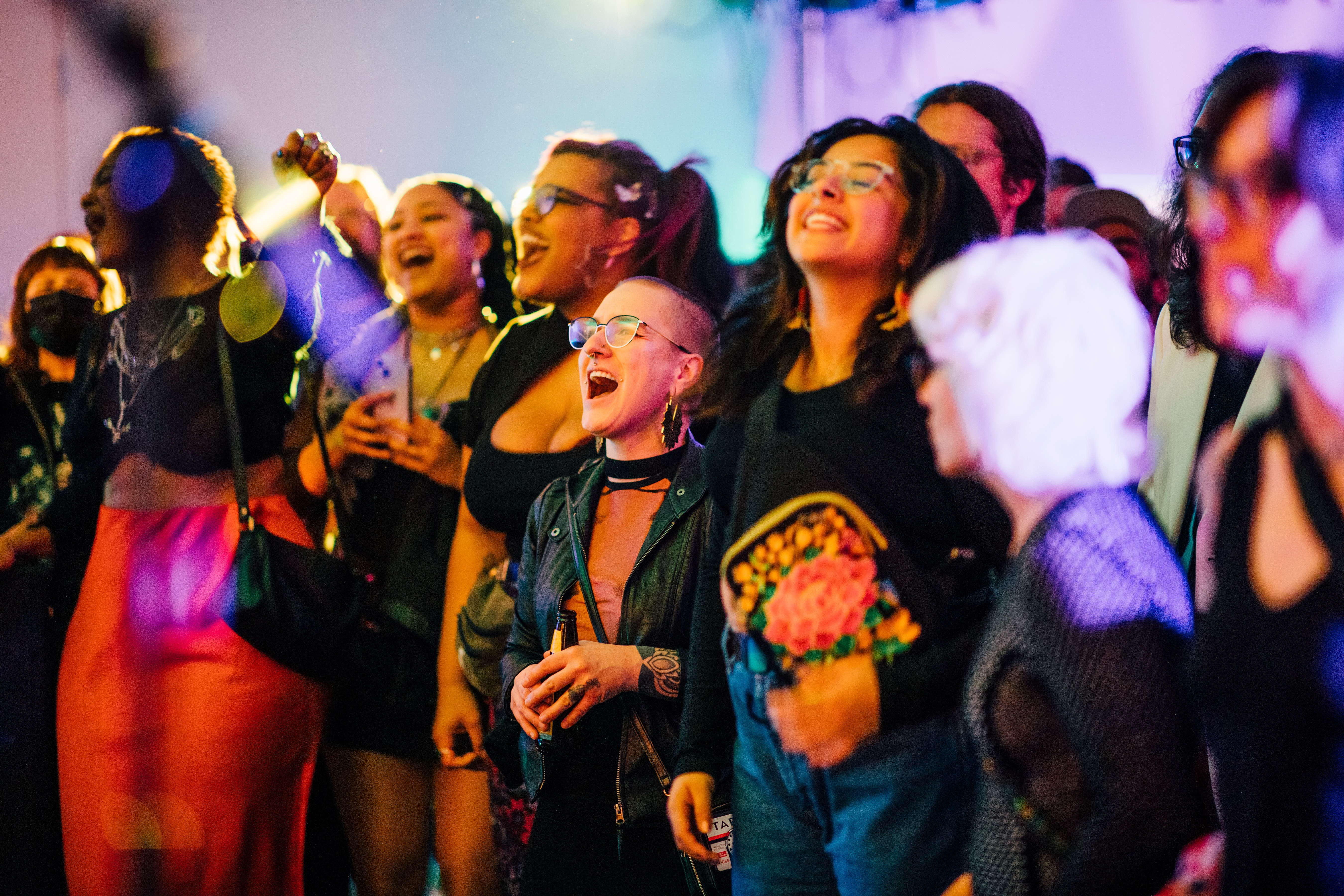 A crowd of people stand together at a brightly lit event, some smiling and others clapping or singing along.