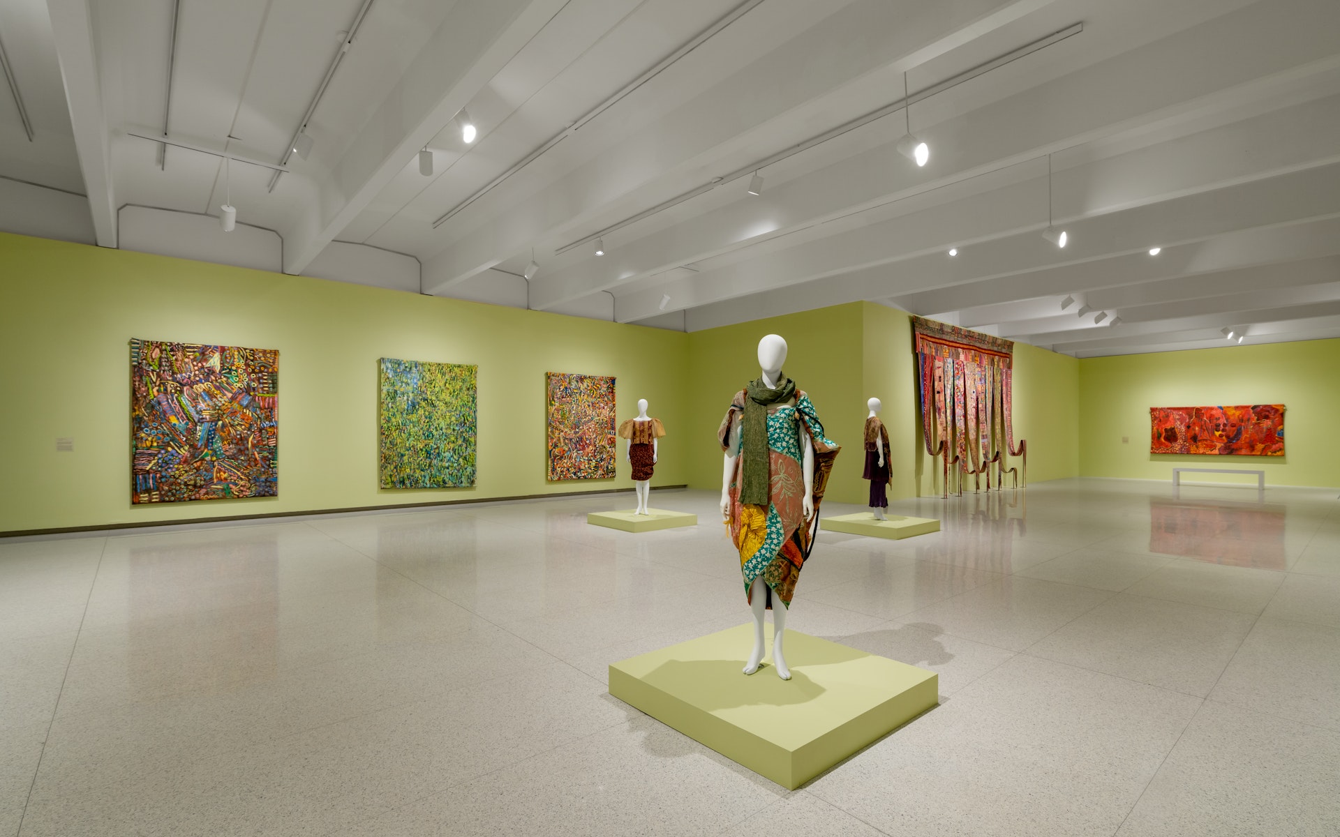 View of a art gallery with green walls that have colorful textile works hanging from the walls and colorful wrap dresses on manniquins.