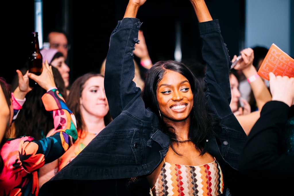 A woman with dark skin smiling with arms raised, surrounded by people dancing and celebrating at a lively event.