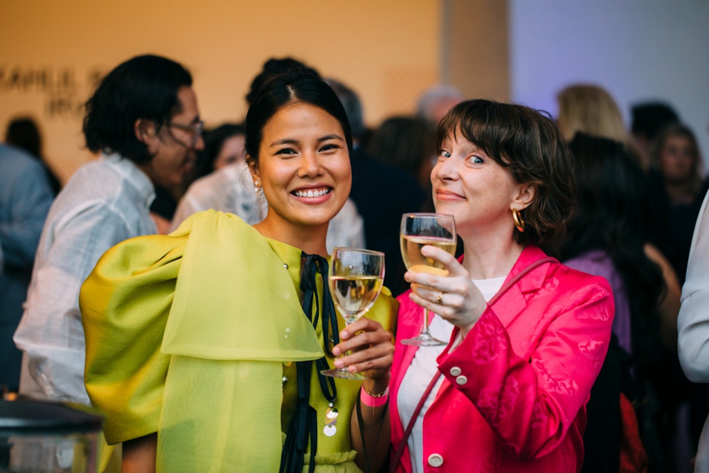 Two adults at a party look at the viewer as they raise glasses of wine towards them.