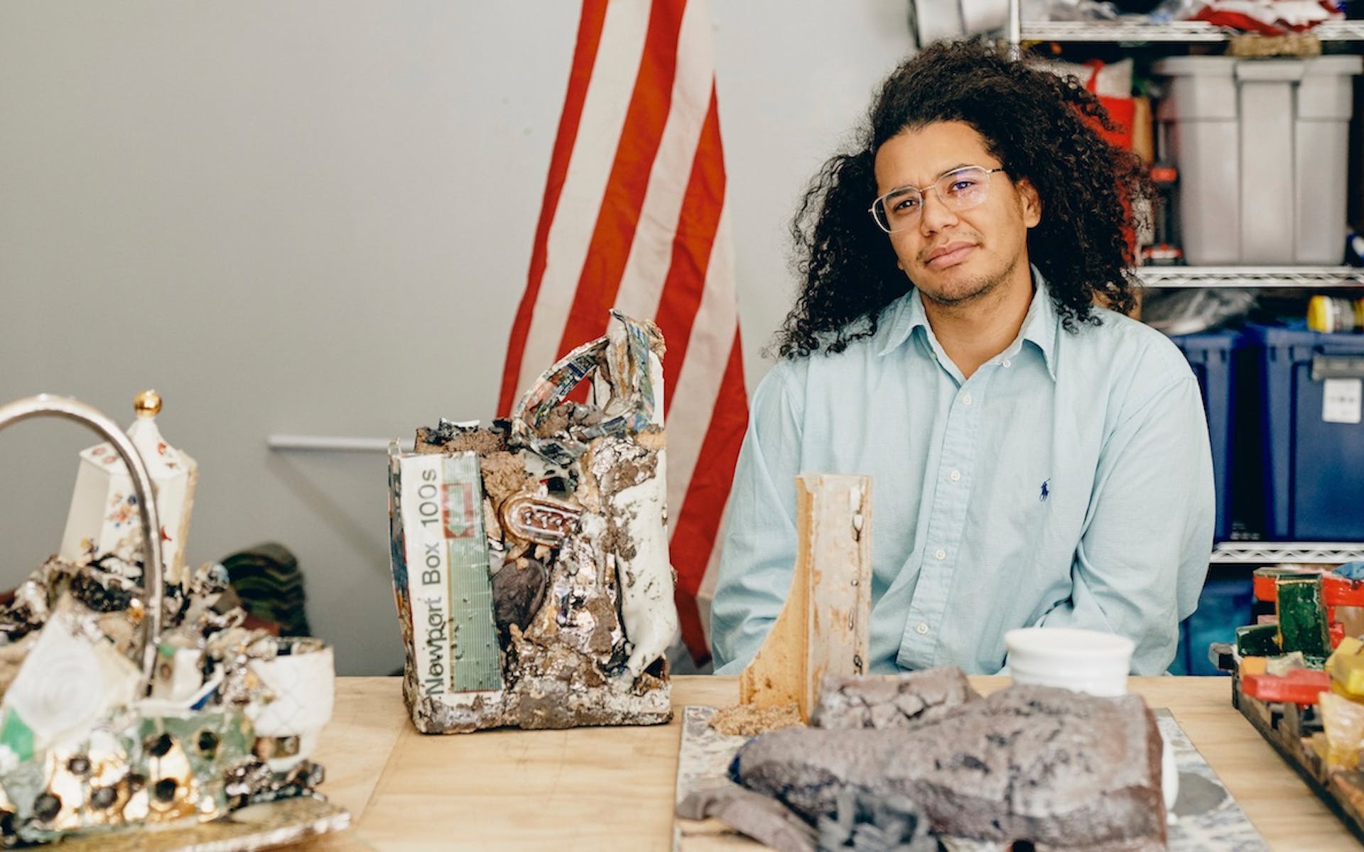 A man sits in a studio at a desk with sculptures made from clay and objects in the street with an American flag on a flagpole behind him.