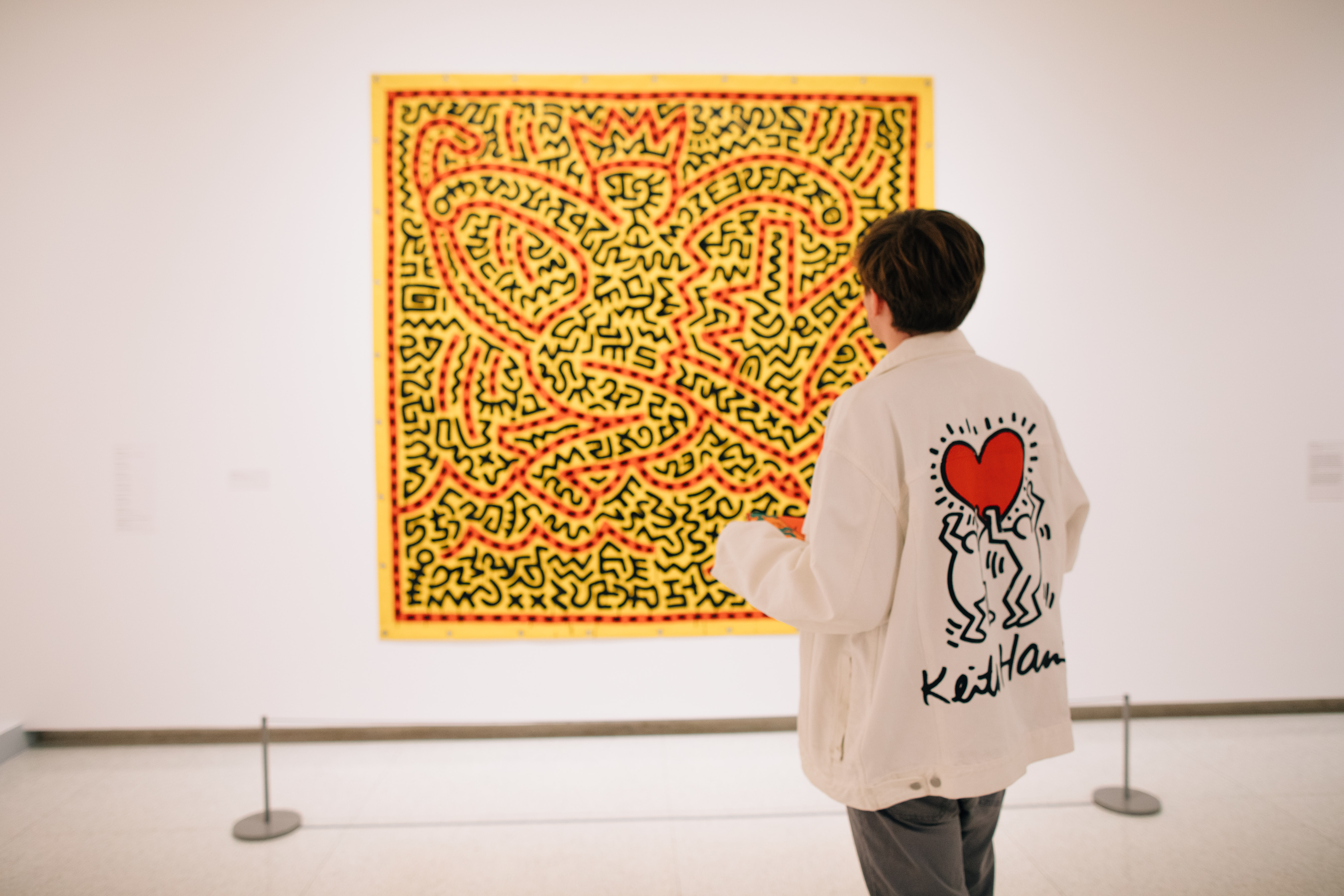 An adult with a white jacket that has Keith Haring art on the back stands with their back to the viewer looking at Keith Haring art on the wall in a gallery.