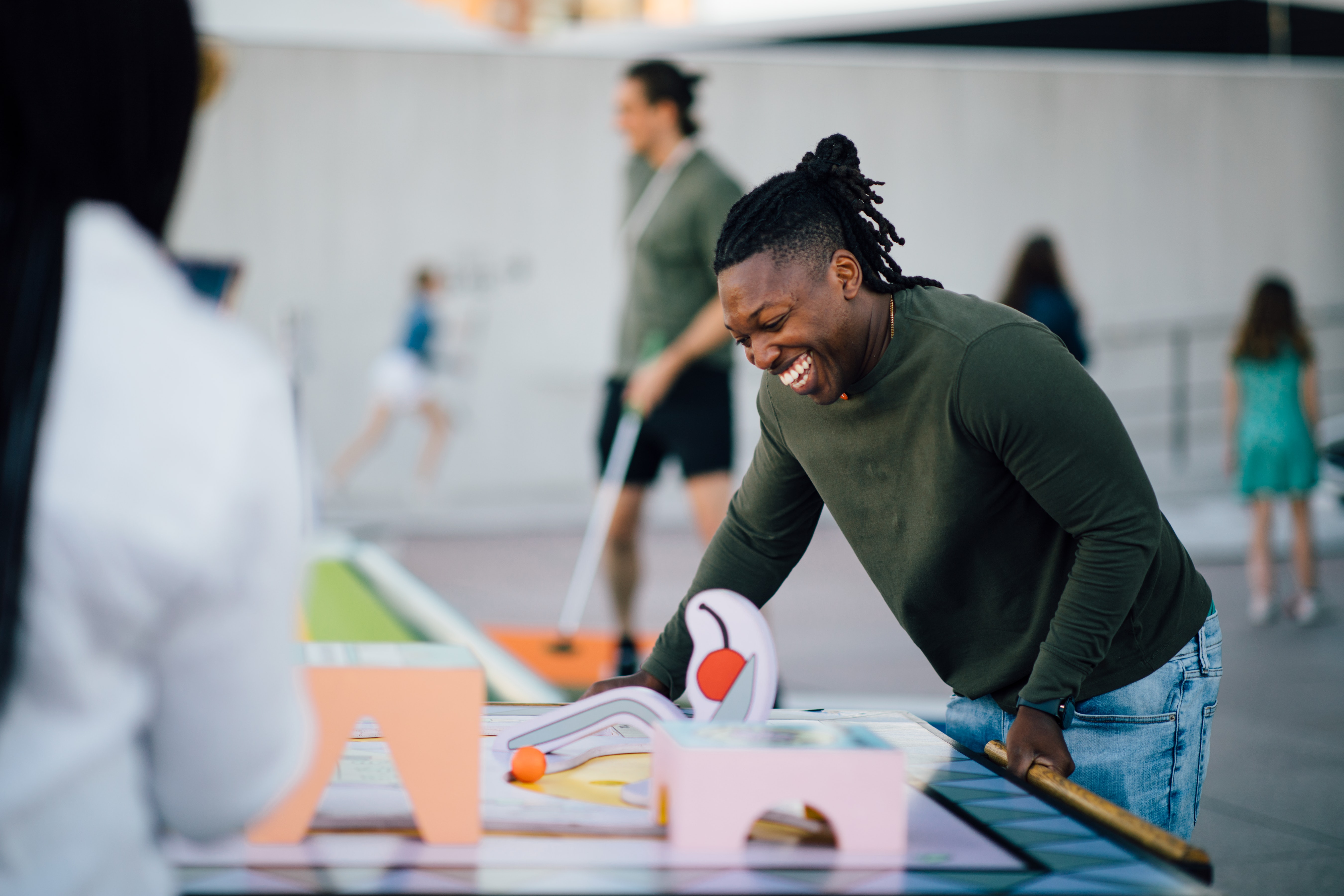 A man with dark skin smiles and plays a colorful tabletop game outdoors, with people in the background.