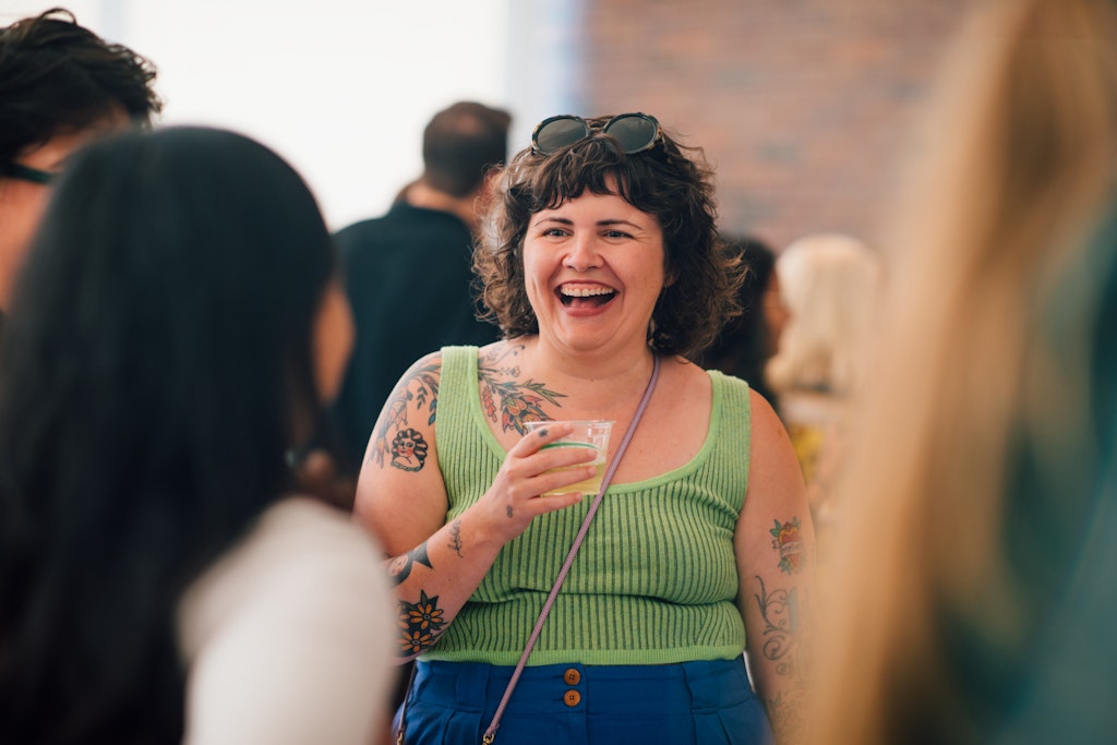 A woman with tattoos smiles and holds a drink while socializing at a lively indoor gathering.