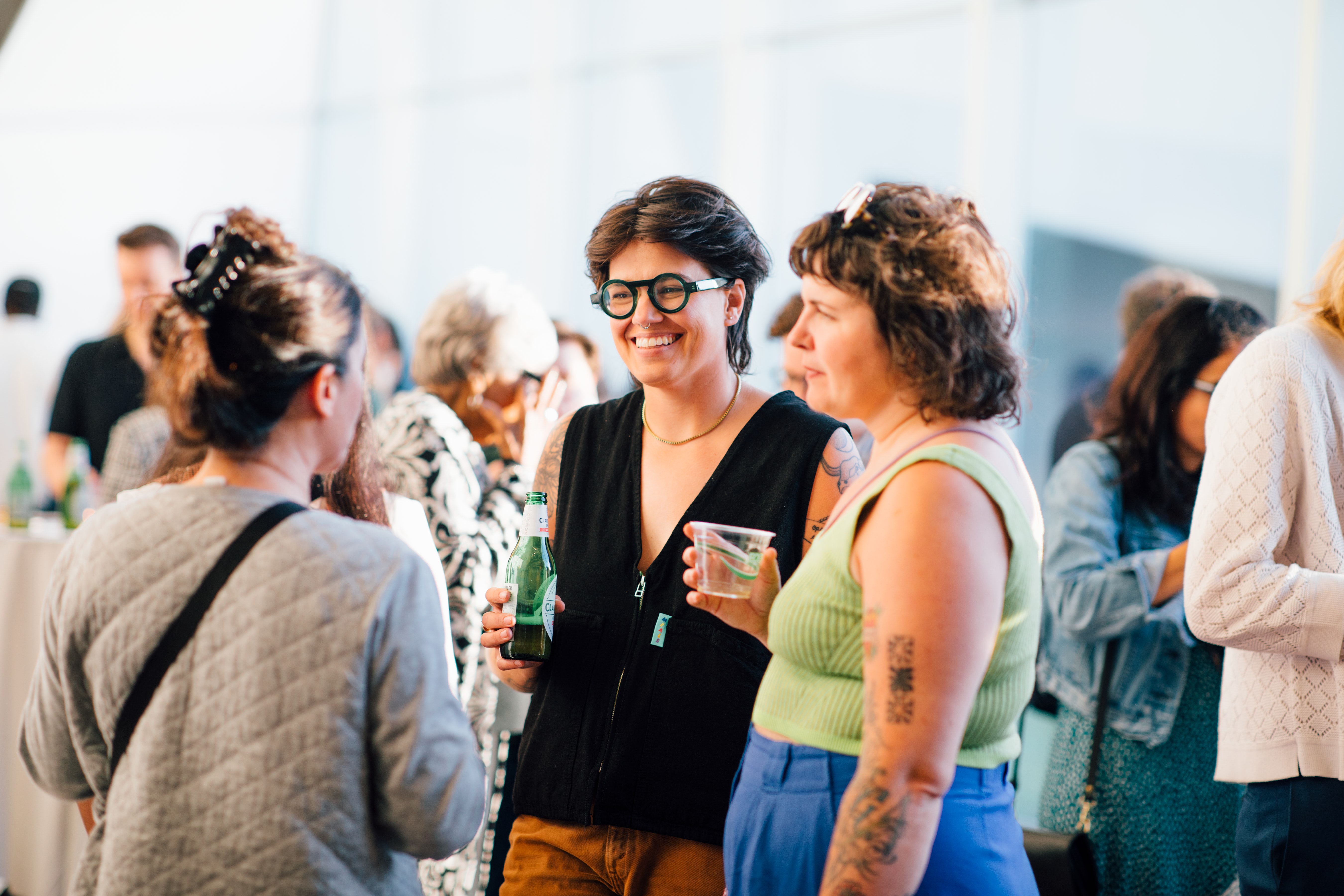 A group of three stand talking during a reception at the Walker. One holds a bottle of beer, another a plastic class of wine.