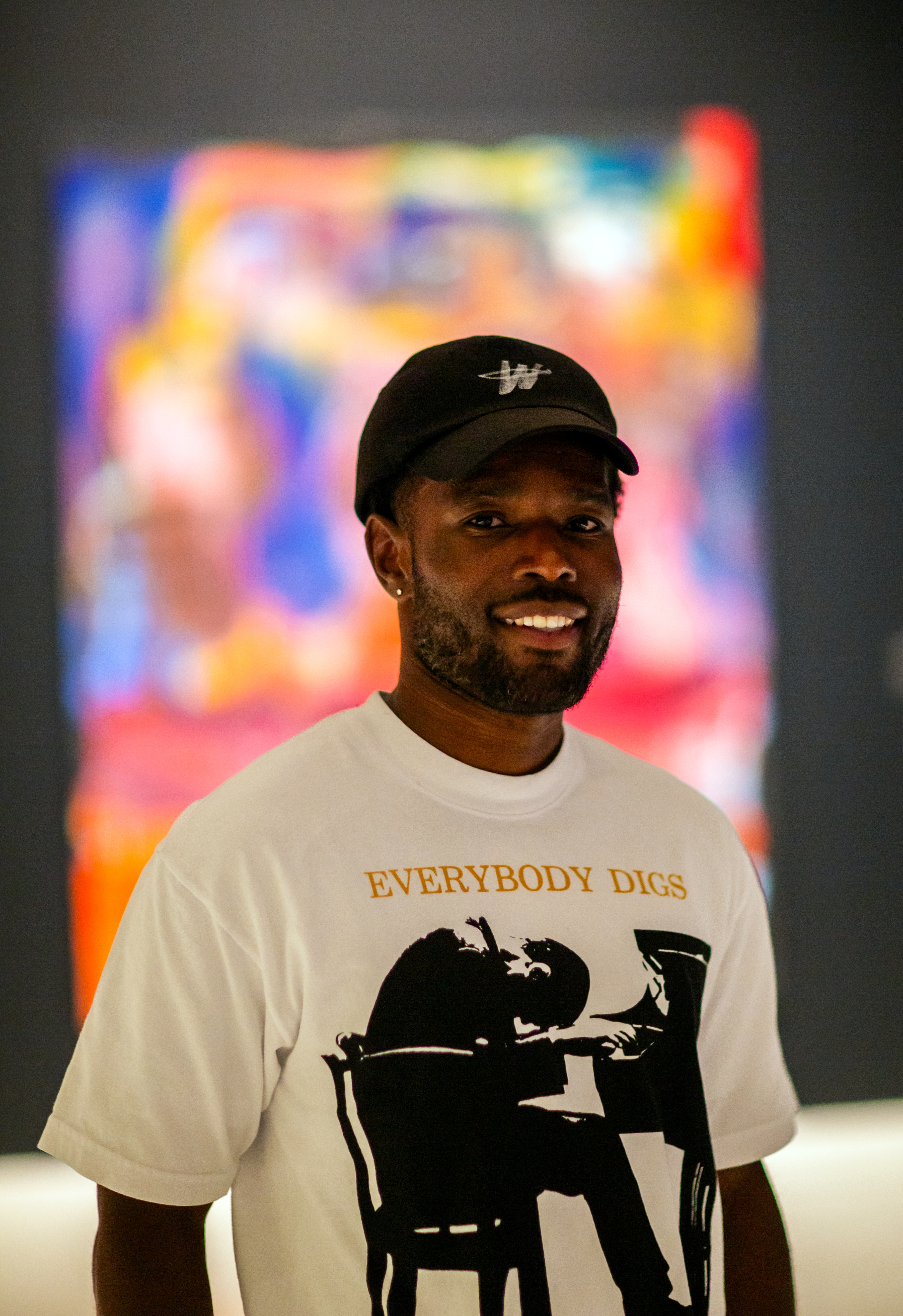 An adult with dark skin, black hat and white t-shirt stands facing the viewer smiling in front of a colorful background