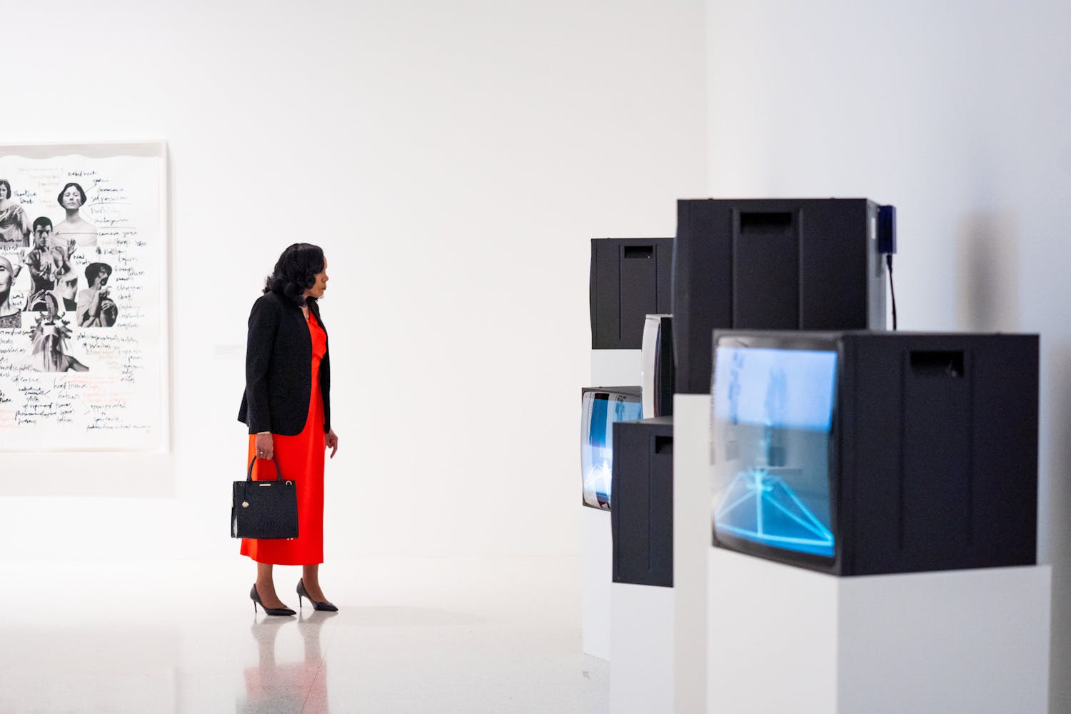 A woman with dark skin holding a purse watches videos on a series on monitors in a white gallery.