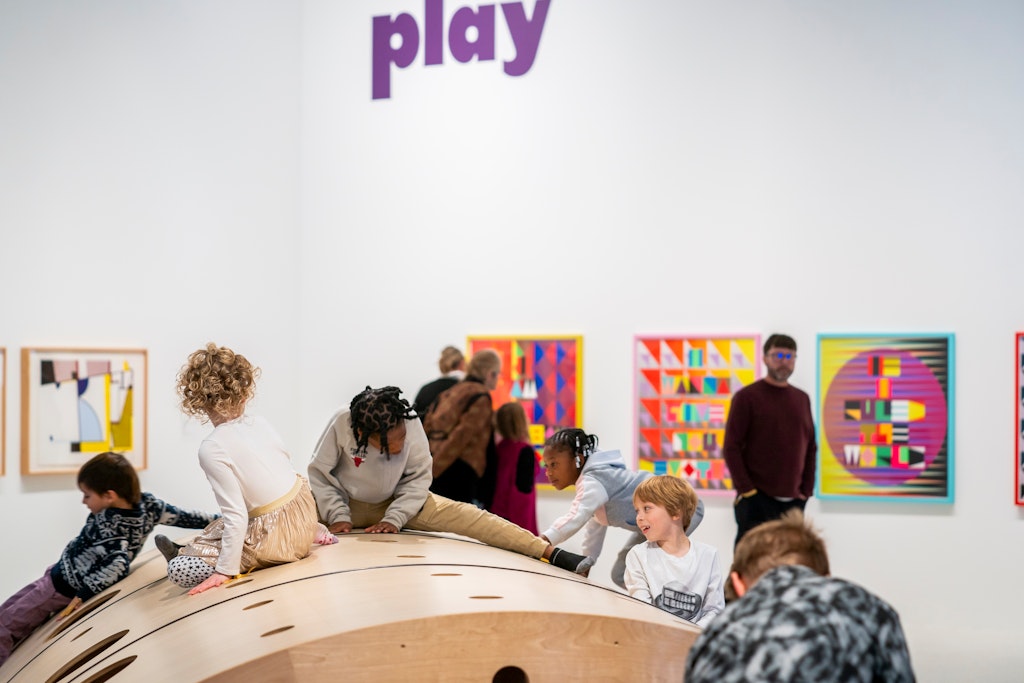 Children climb and play on a large wooden interactive structure inside a bright art gallery, while colorful geometric artworks hang on the walls behind them. Several adults observe in the background, and the word “play” appears in large purple letters on the wall above.