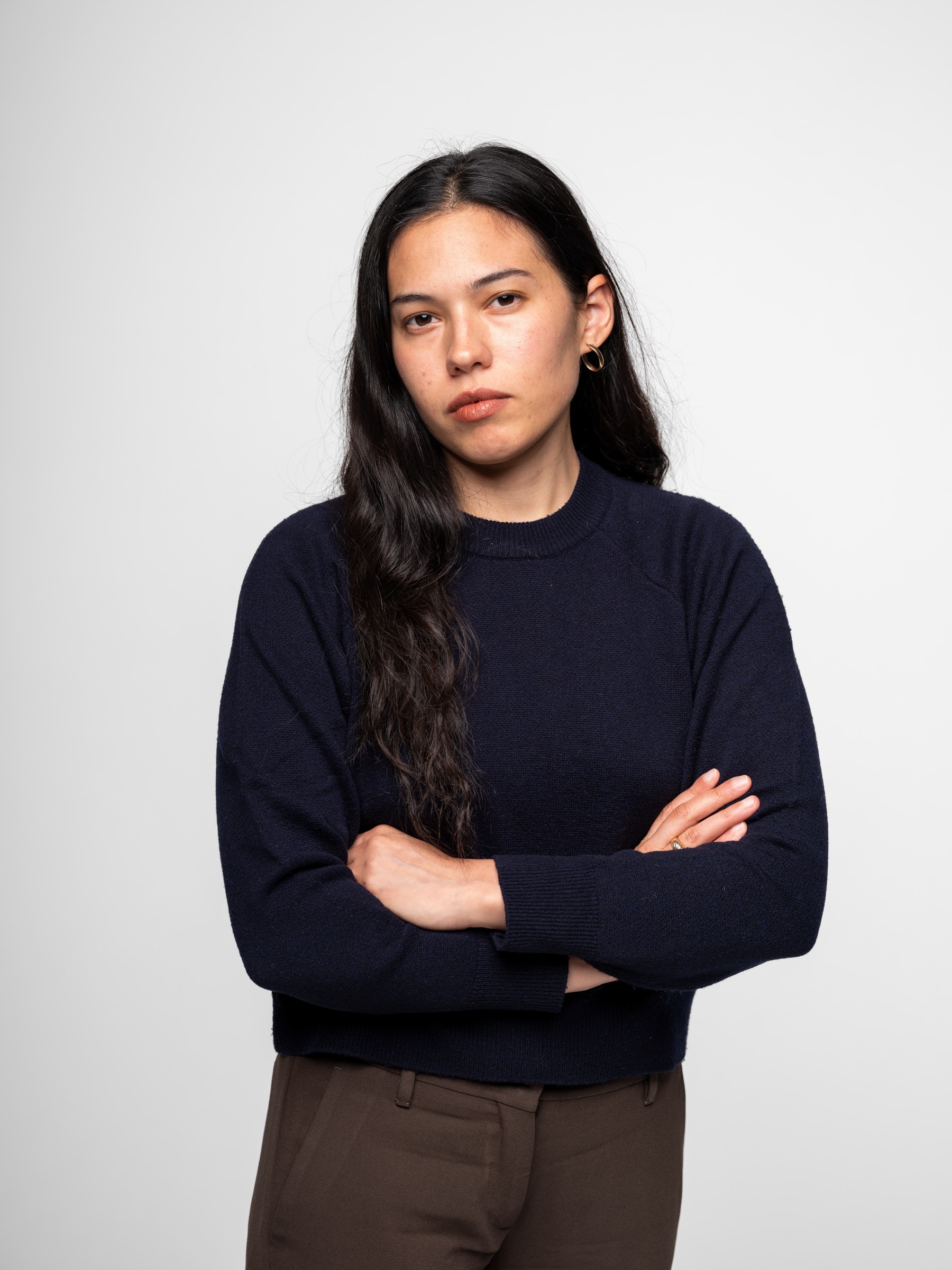 Woman with long dark hair wearing a navy sweater stands with arms crossed against a plain background.