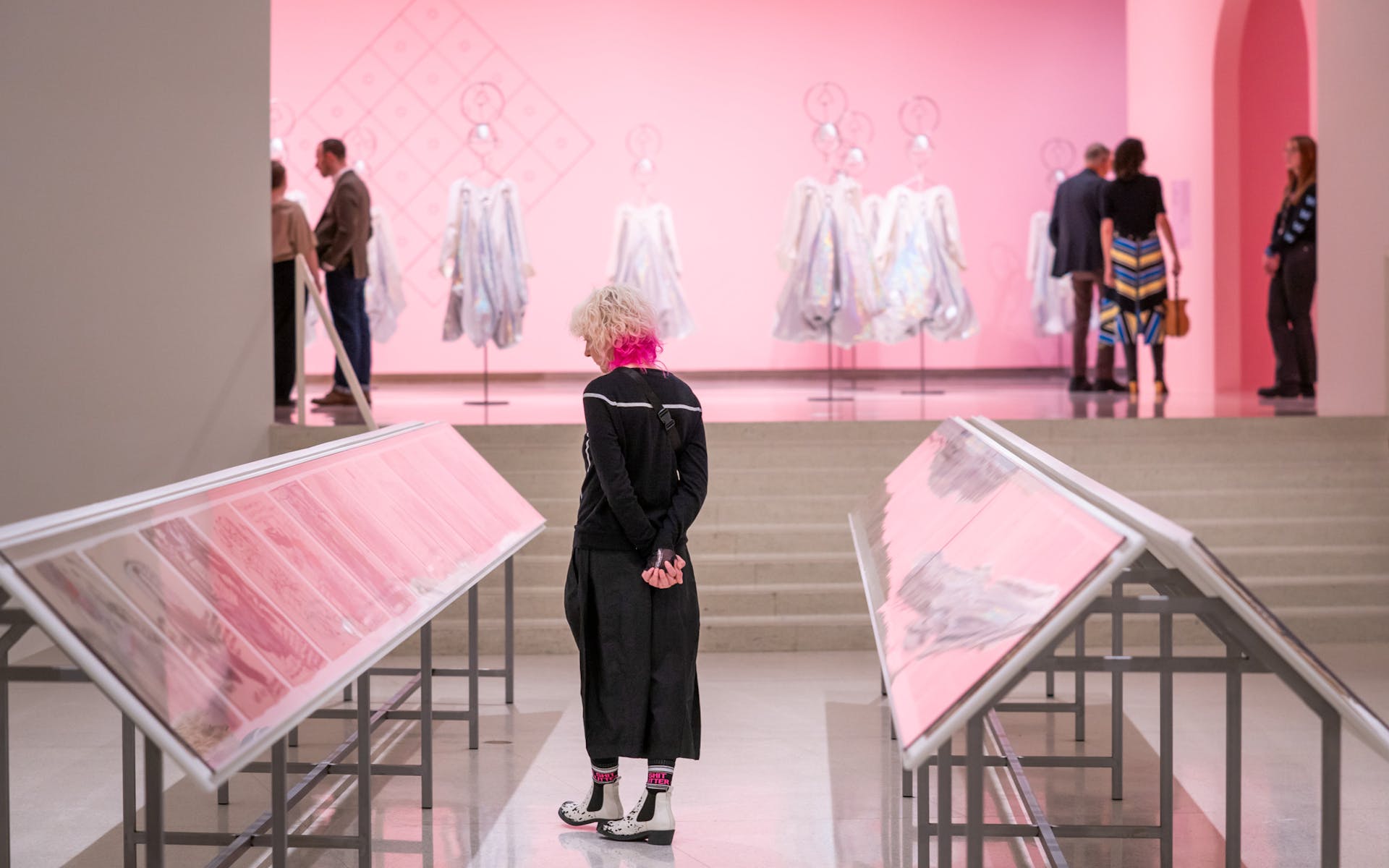 Person with white hair stands in an art gallery with pink walls, viewing displays of artwork.