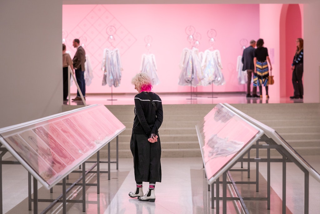 Person with white hair stands in an art gallery with pink walls, viewing displays of artwork.