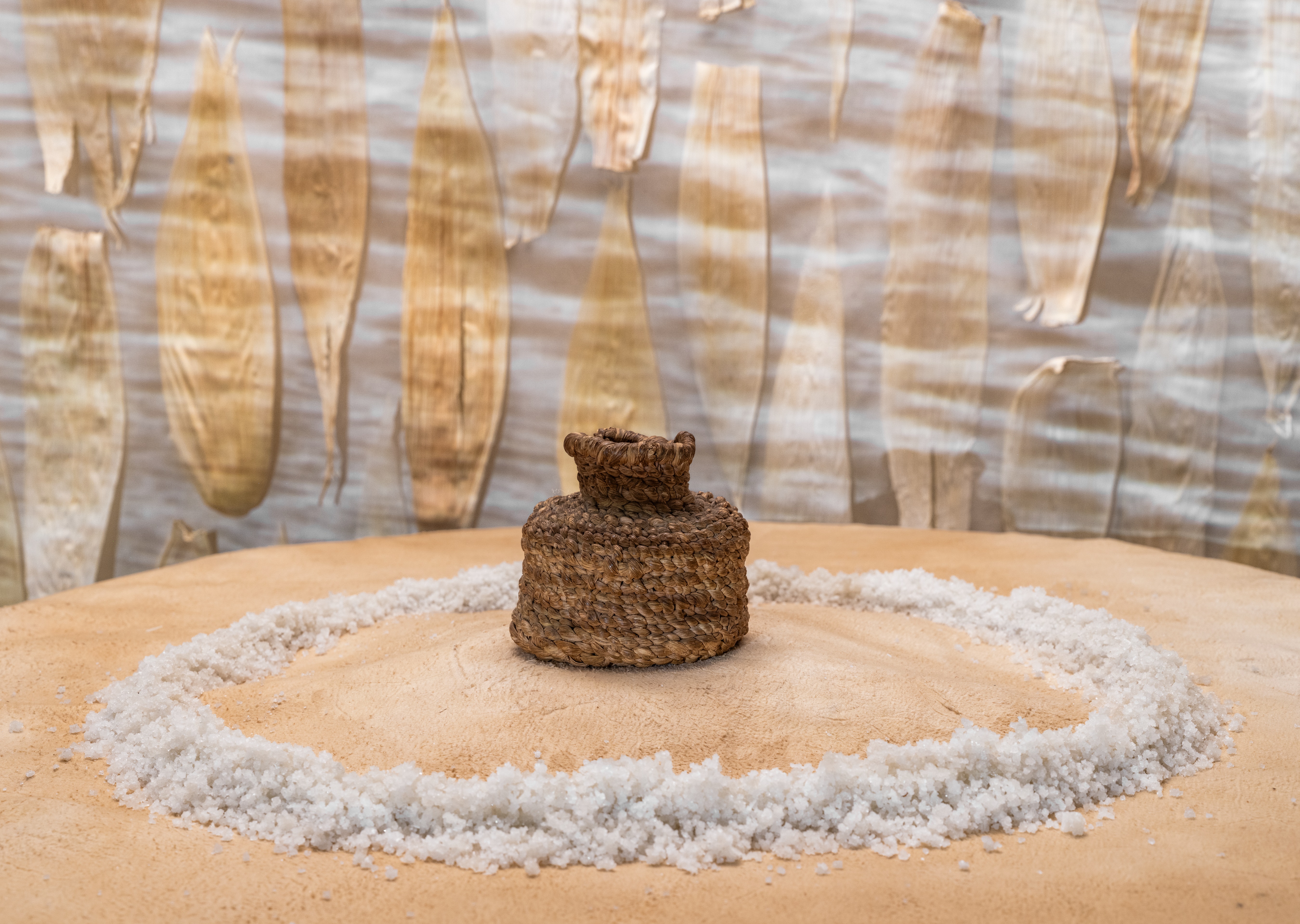 A small brown woven vessel is displayed on a tanned hide, surrounded by a ring of coarse white salt. In the background, a black & white video still of rippling water projects onto vertical rows of corn husks.
