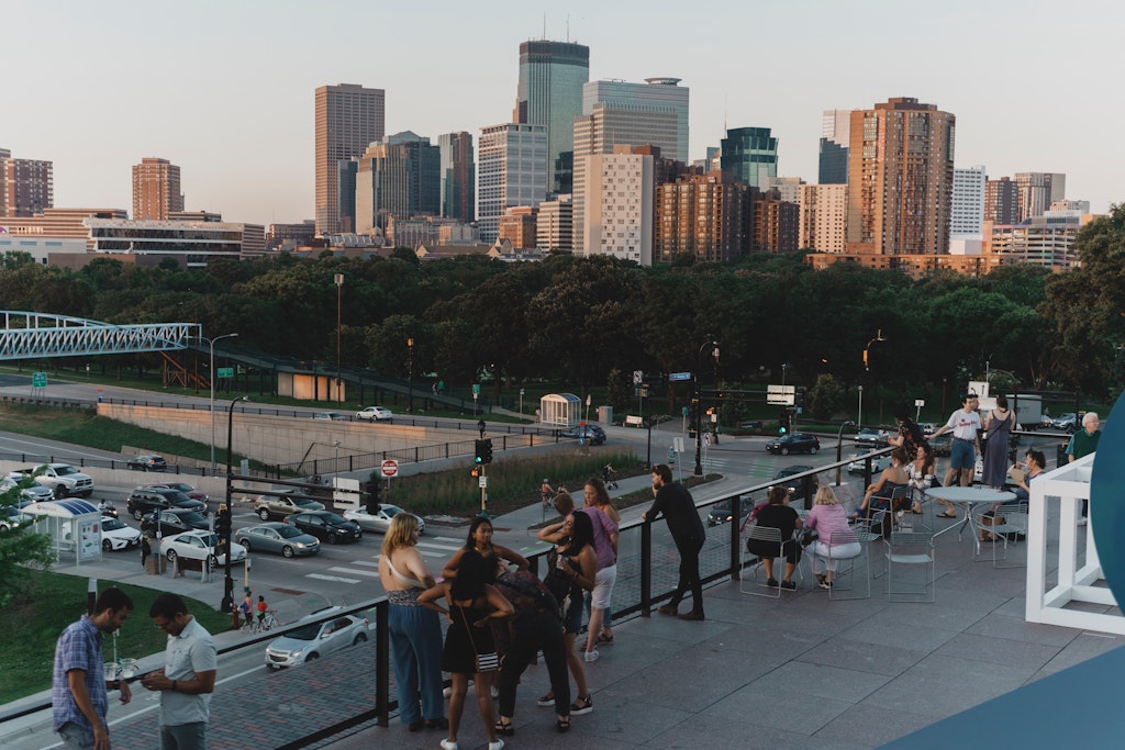 People gather on a terrace overlooking a busy street, with a city skyline in the background at sunset.