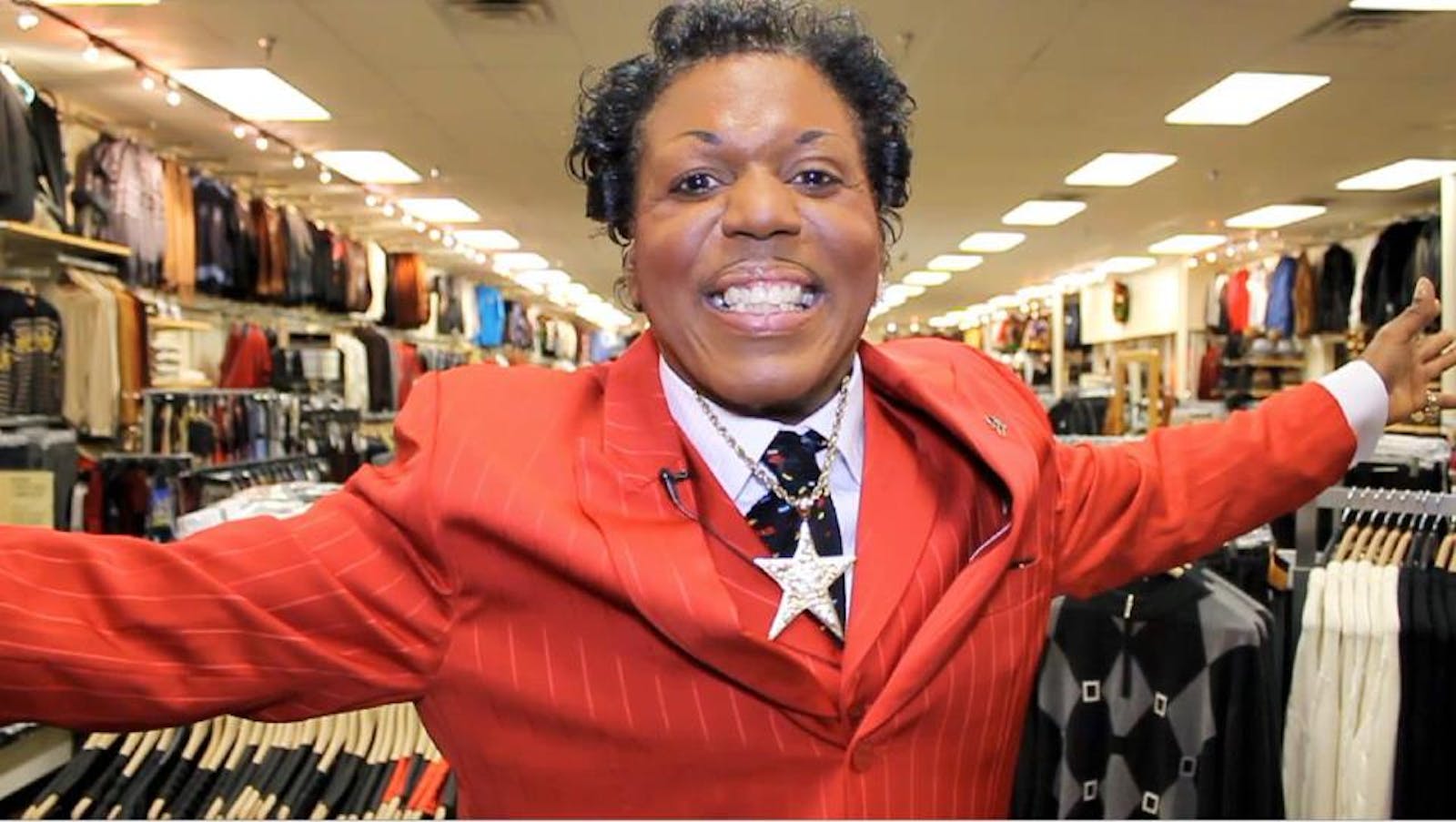 A man in a red suite holds his arms outstretched and smiles at the viewer.