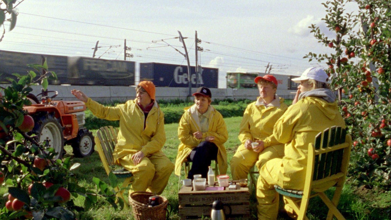 Four people sit in yellow rain gear on a lawn eating and drinking while a train of shipping containers passes behind them.