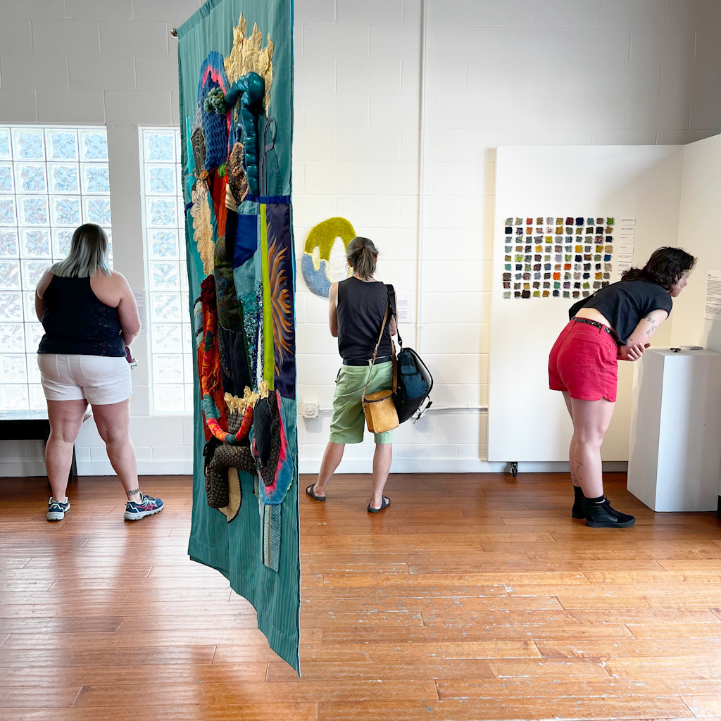A group of adults look at craft artworks on display in a white gallery.
