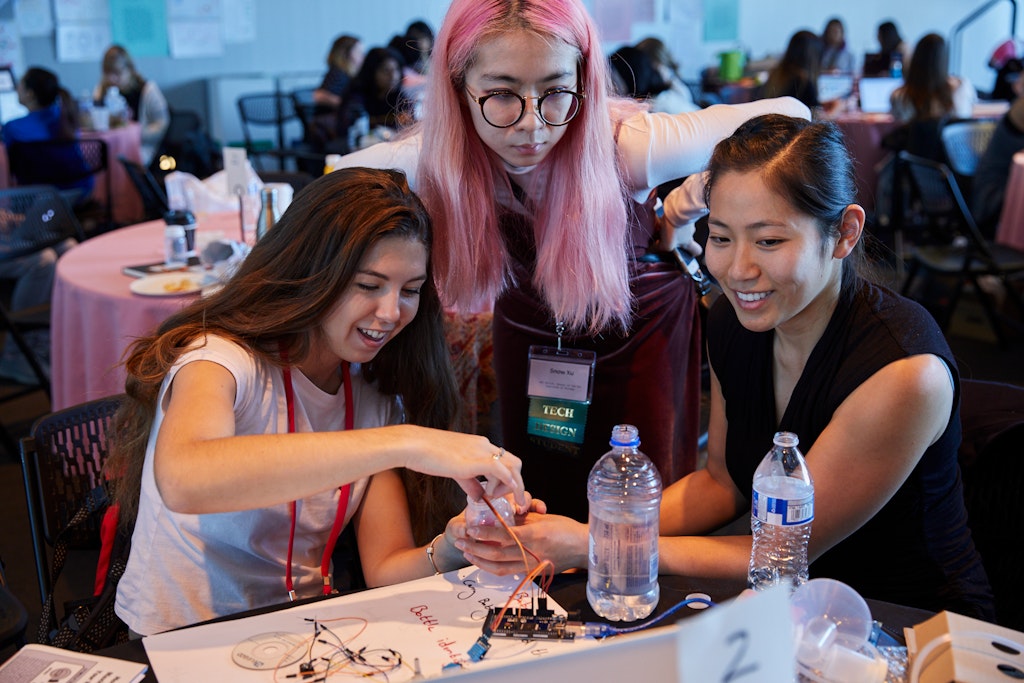 Three young women sitting at a table together working on what appears to be a circuit board.
