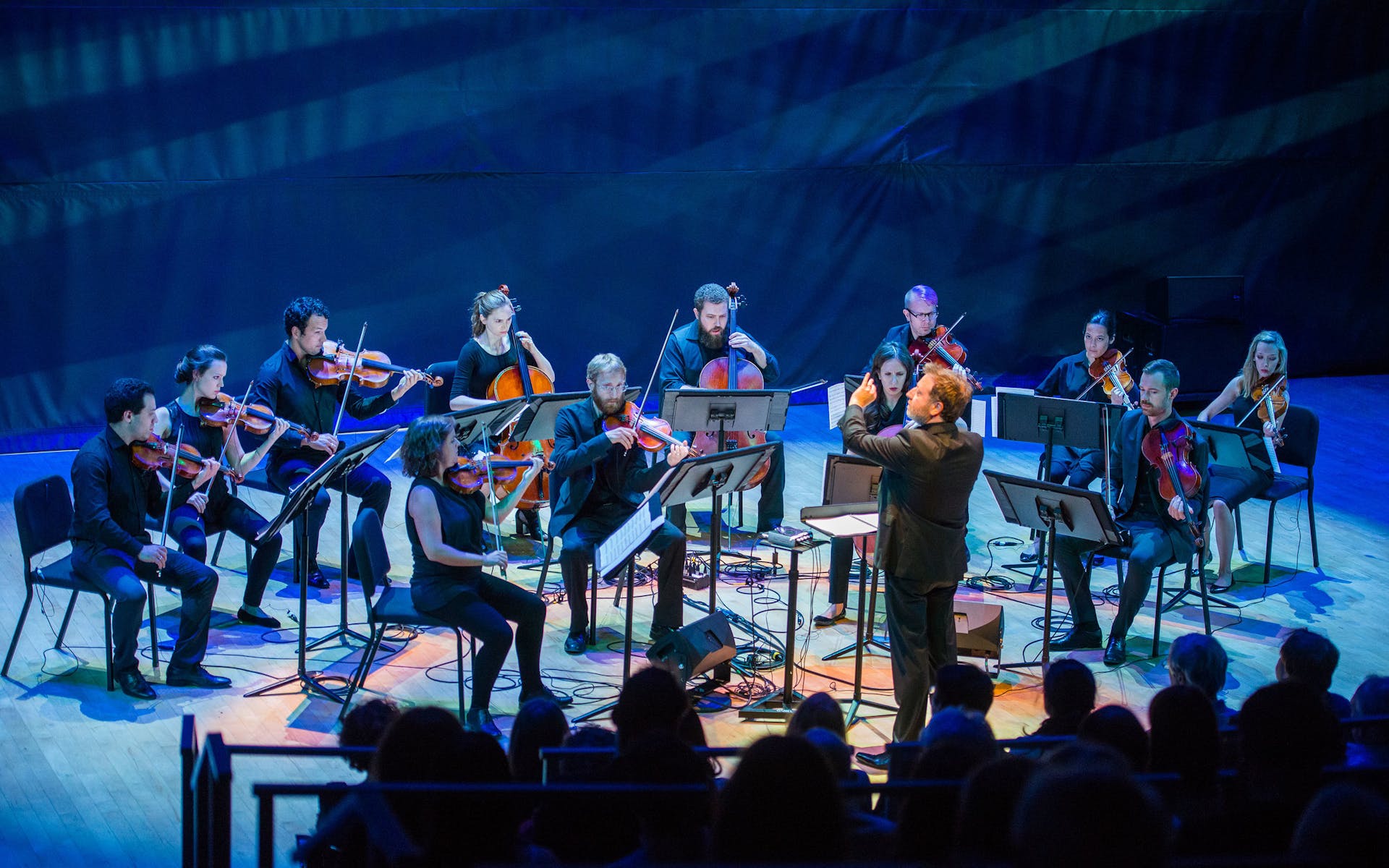 A strings ensemble plays music in front of a conductor while bathed in projected blue lights.