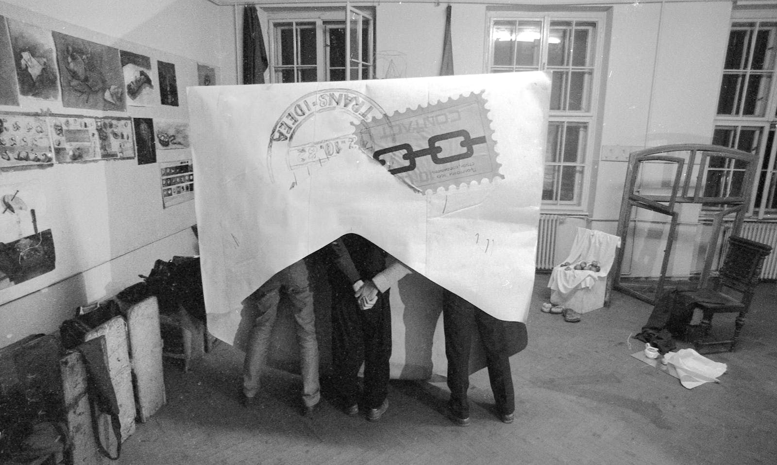 Three adults hold hands while the stand with a giant envalope over their heads and torosos in a studio.