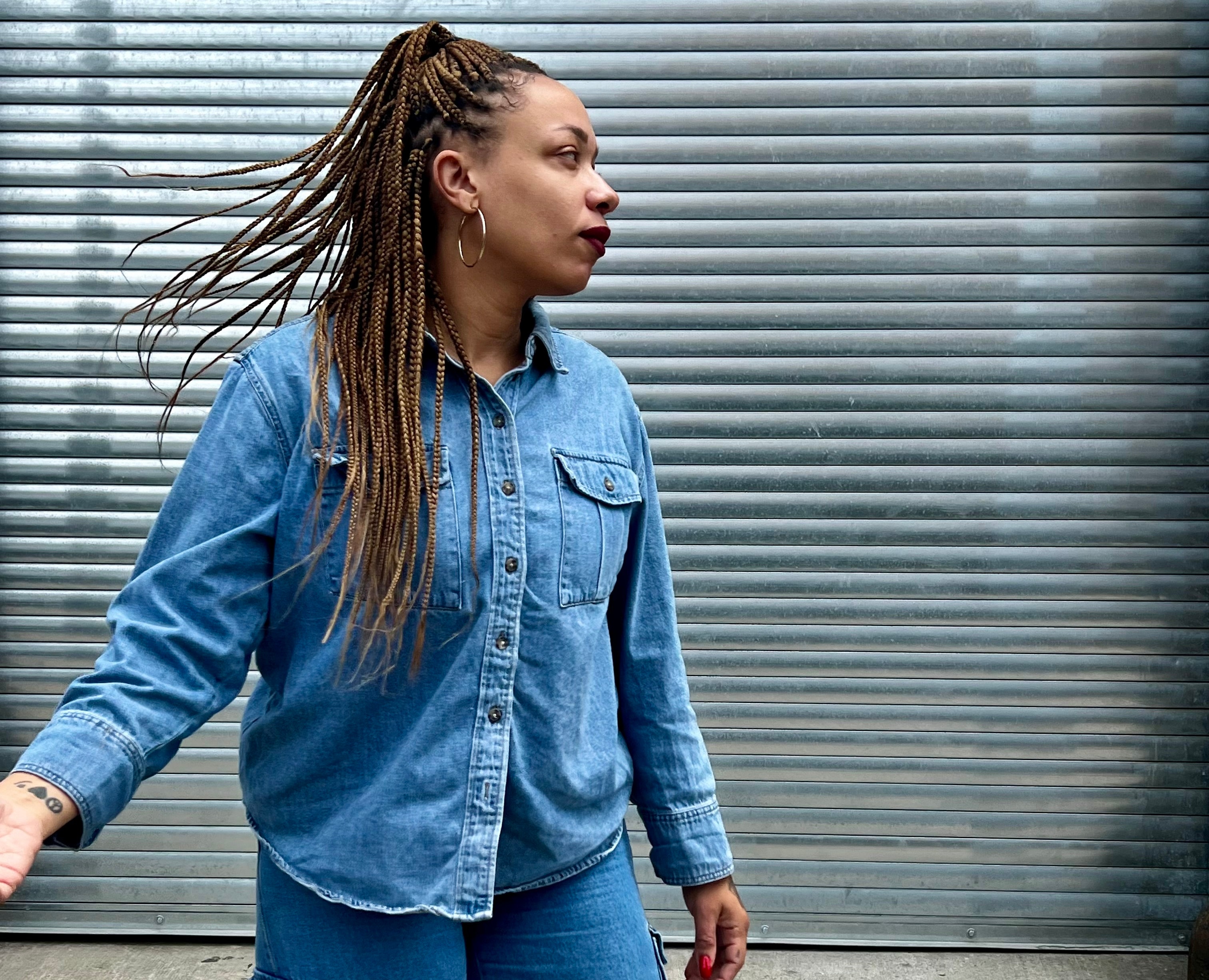 An adult with medium dark skin and long cornrows, wearing all denim is in action looking to the side