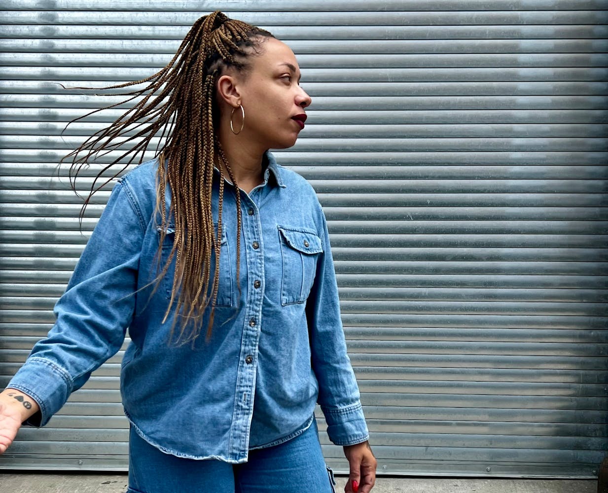 An adult with medium dark skin and long cornrows, waering all denim is in action looking to the side