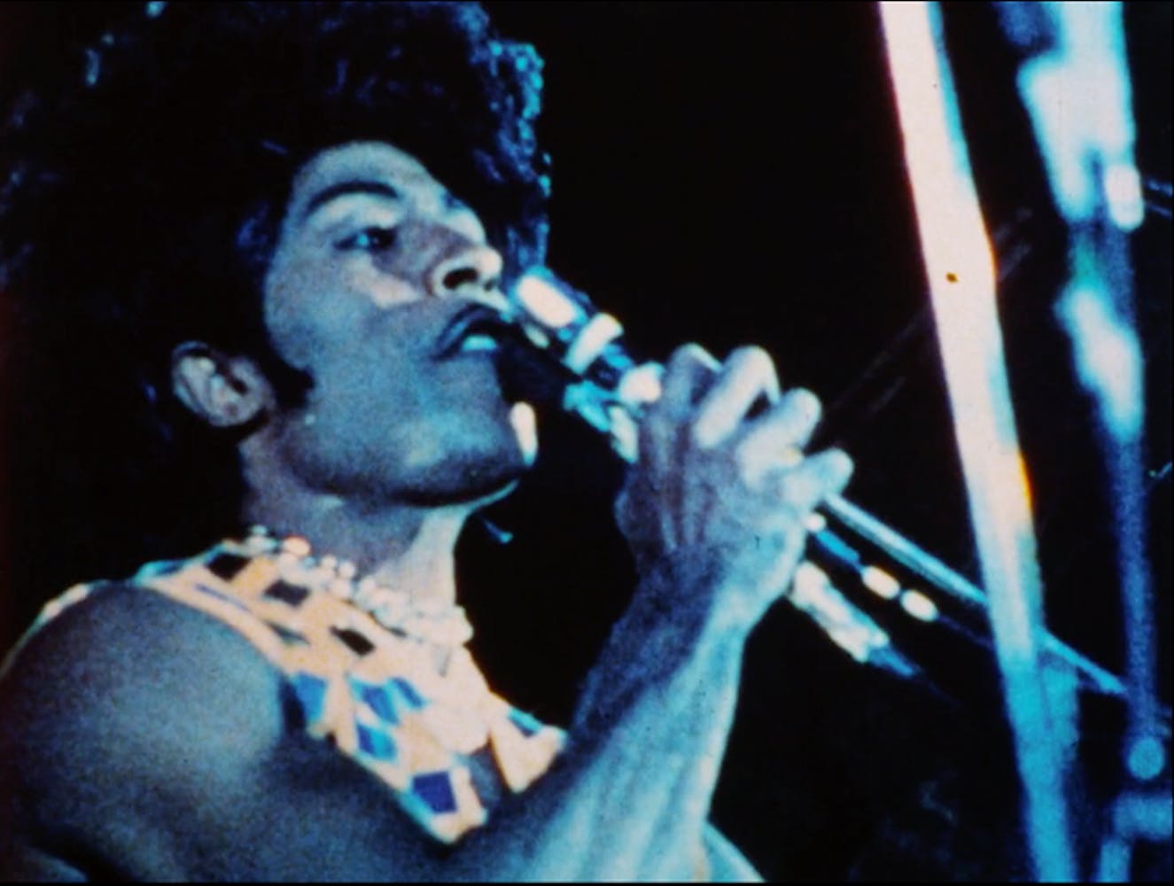 Little Richard performing under blue stage lights. He wears a pompadour, a mirrored shirt and holds a microphone to his mouth.