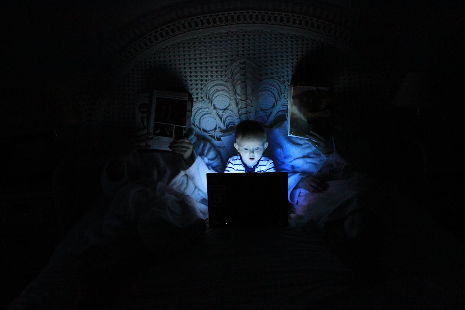 A baby watches a laptop in a dark bedroom while sitting between slepping parents.