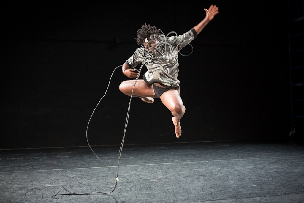 A performer with dark skin wrapped in microphone cabling jumps with left arm outstretched.