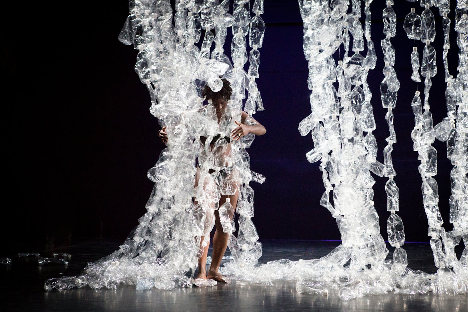 A performer with dark skin grabs strings of plastic bottles suspended from above.
