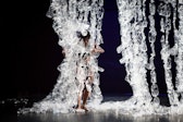 A performer with dark skin grabs strings of plastic bottles suspended from above.