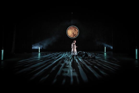 A man walks across a dark stage underneath a large broze colored circle.