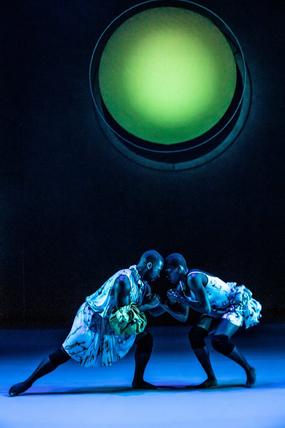 Two adults on stage look arms while facing one another under a large green circle.