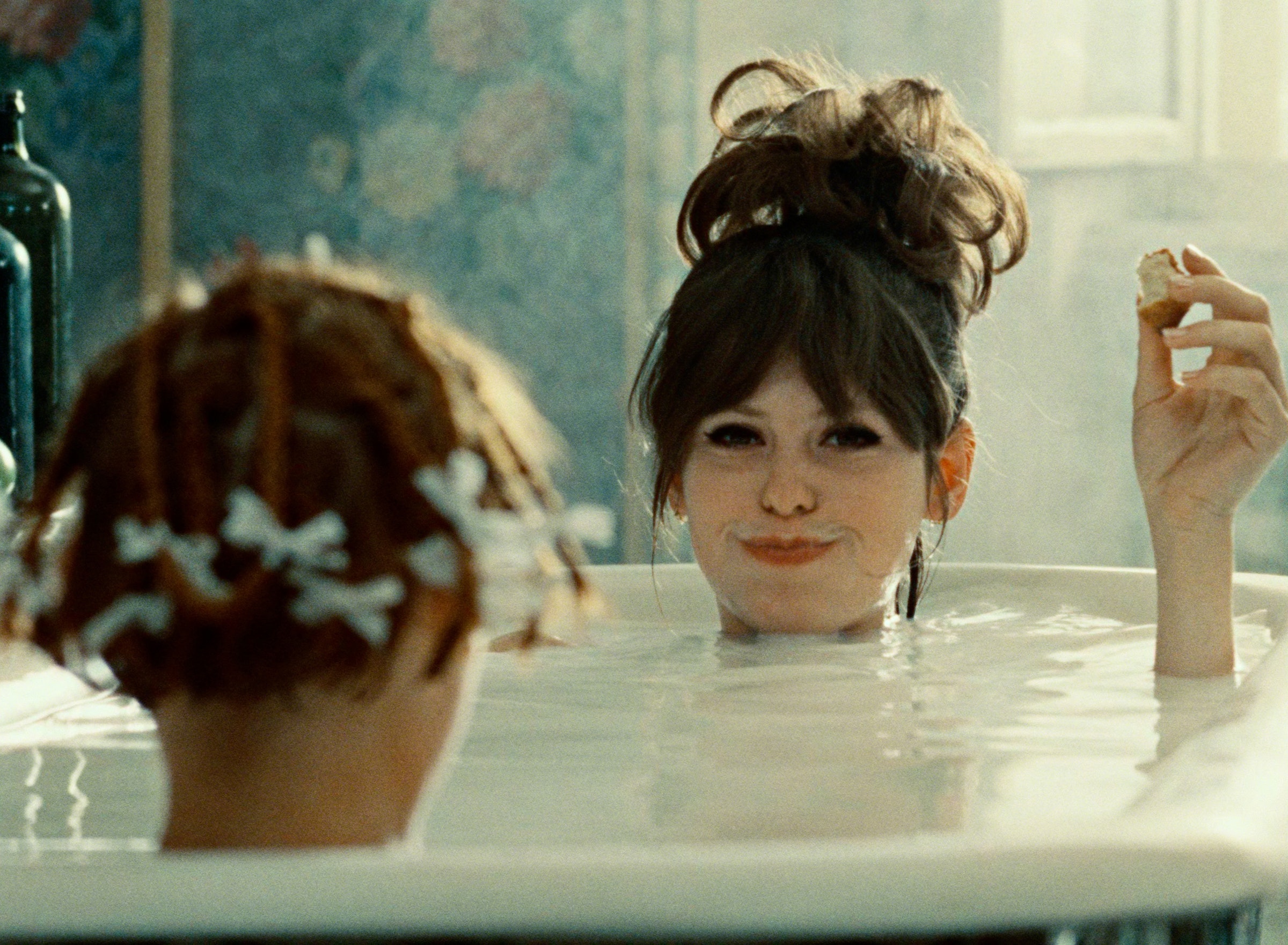 The back of a woman’s head with short hair tied in twists, and another woman’s face smirking at the camera, both are in a bath.