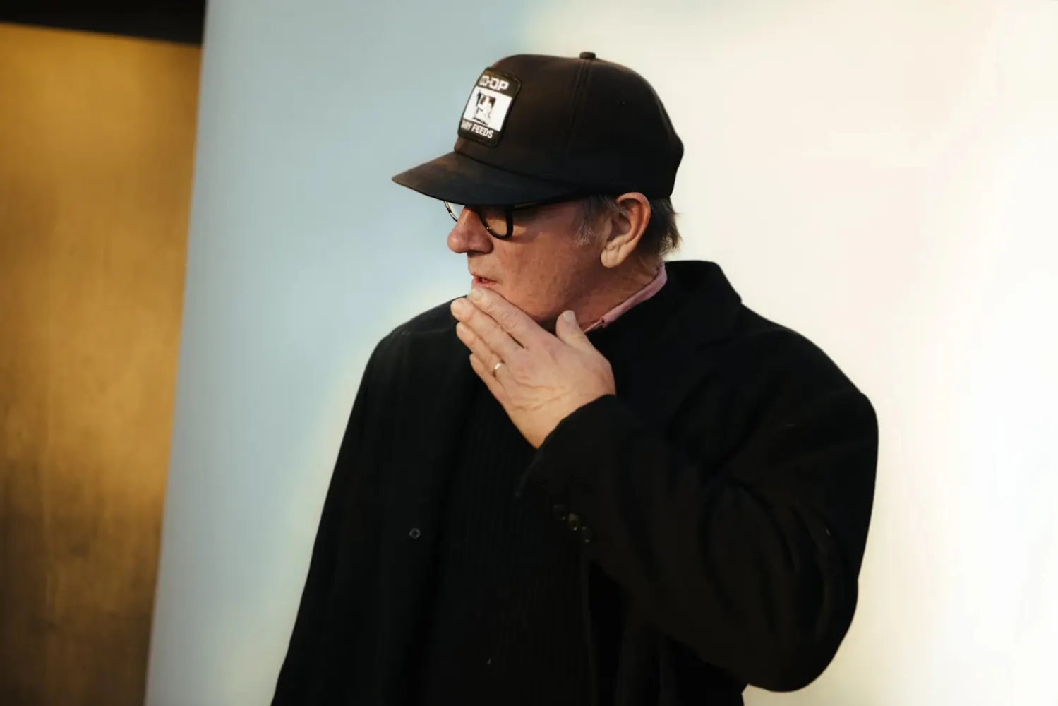 A man in a baseball cap and black suitecoat looks to the side and holds a hand ot his chin.