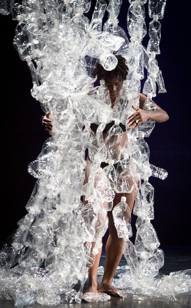 A performer with dark skin grabs strings of plastic bottles suspended from above.