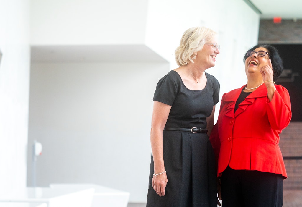 Two women stand next to one another laughing in a hallway of a museum.
