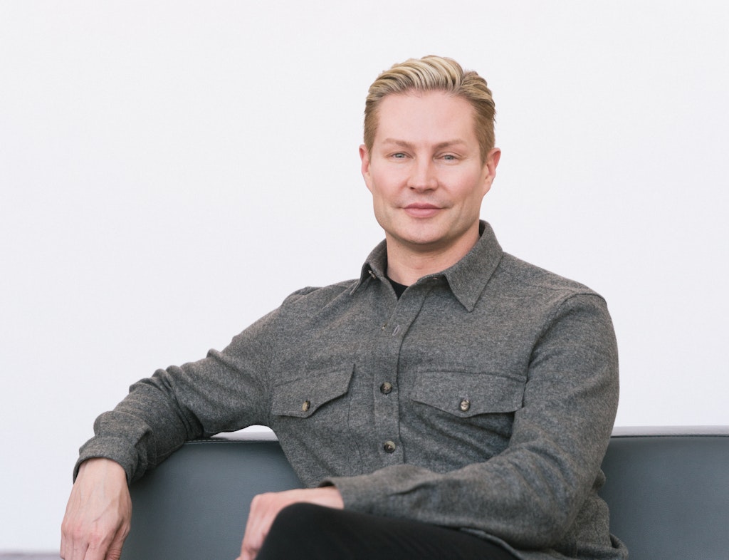 A man sits on a grey couch against a white background and smiles at the viewer.